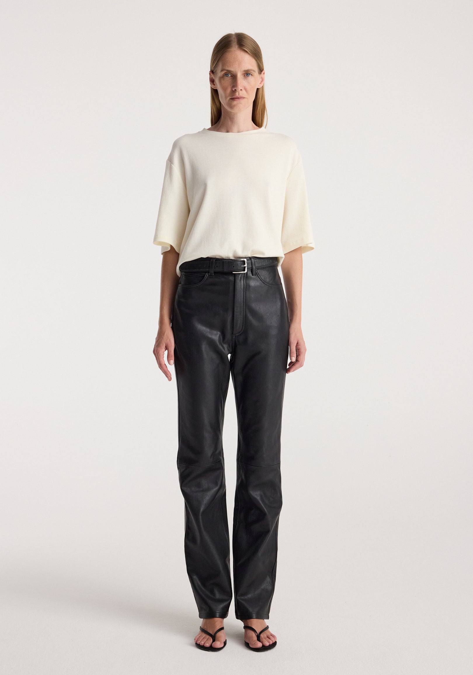 Woman wearing an oversized cotton wool t-shirt | cream paired with high-waisted black leather pants and black open-toe sandals against a plain background.
