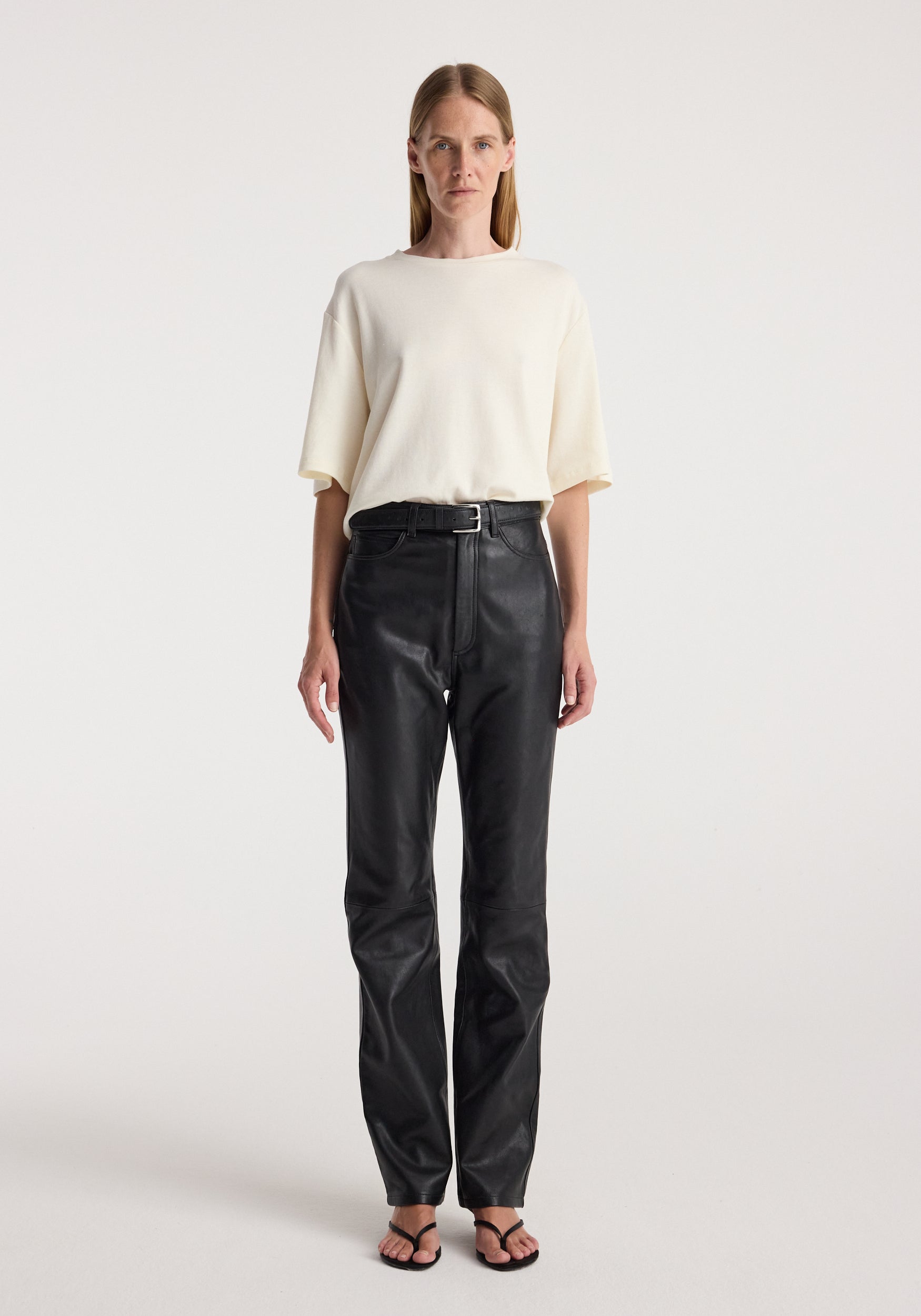 Woman wearing an oversized cotton wool t-shirt | cream paired with high-waisted black leather pants and black open-toe sandals against a plain background.