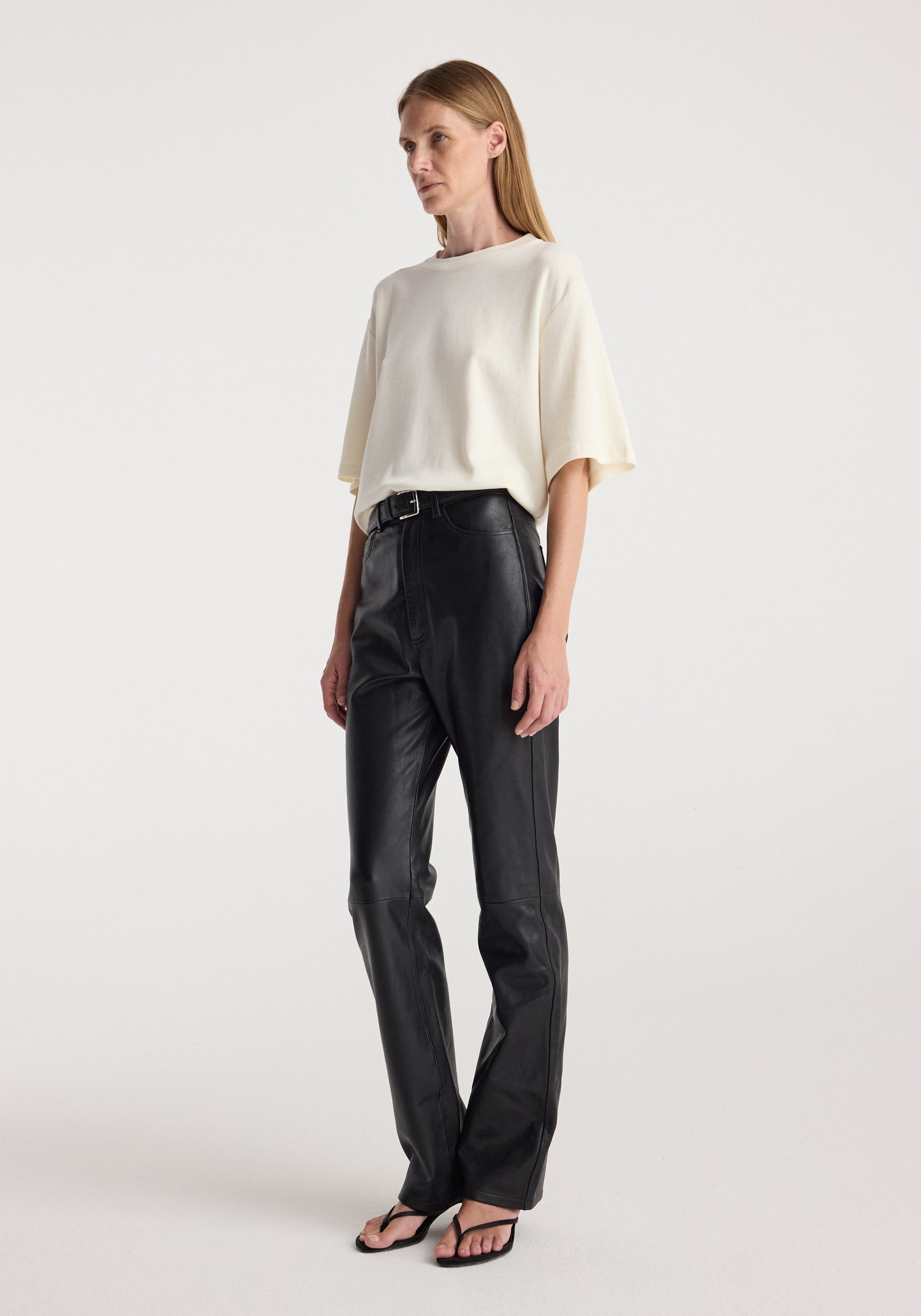 A woman wearing an oversized cotton wool t-shirt | cream with loose short sleeves, paired with high-waisted black leather pants and black open-toe sandals, standing against a plain background.