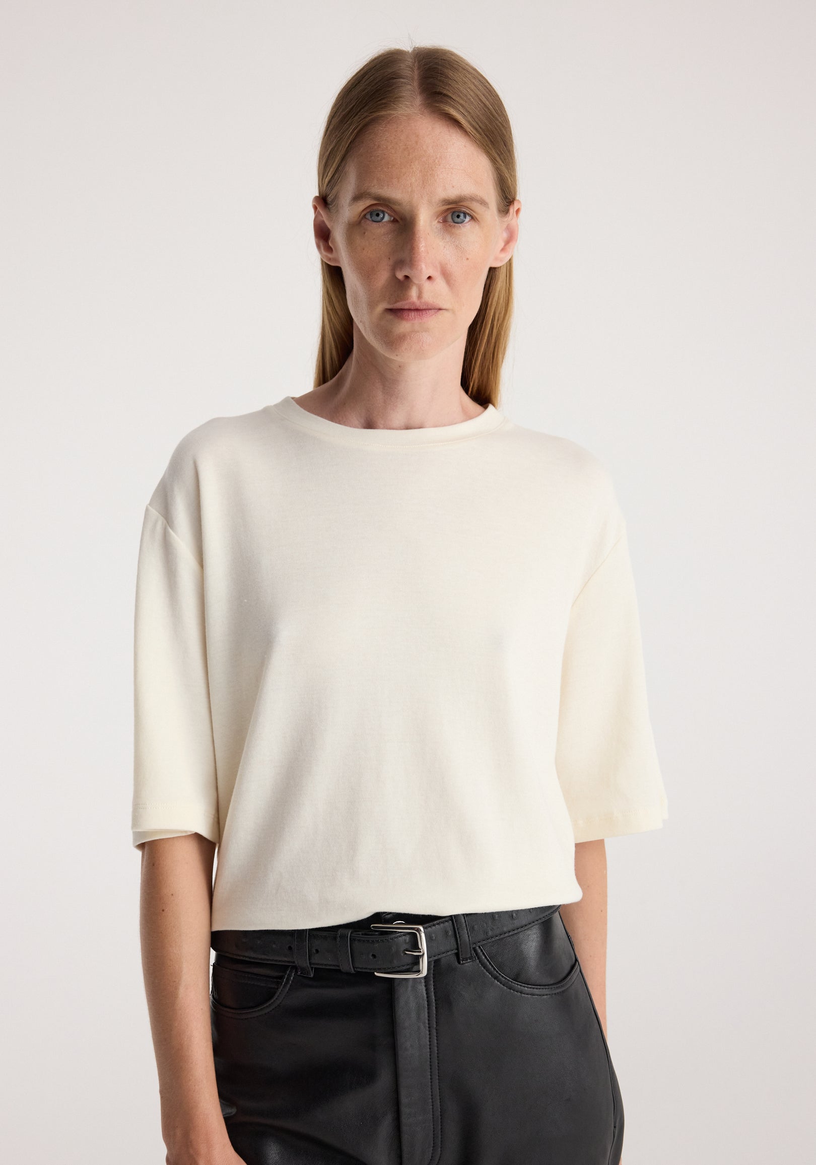 Woman wearing an oversized cotton wool t-shirt in cream, styled with black high-waisted pants and a black belt, standing against a plain light background.