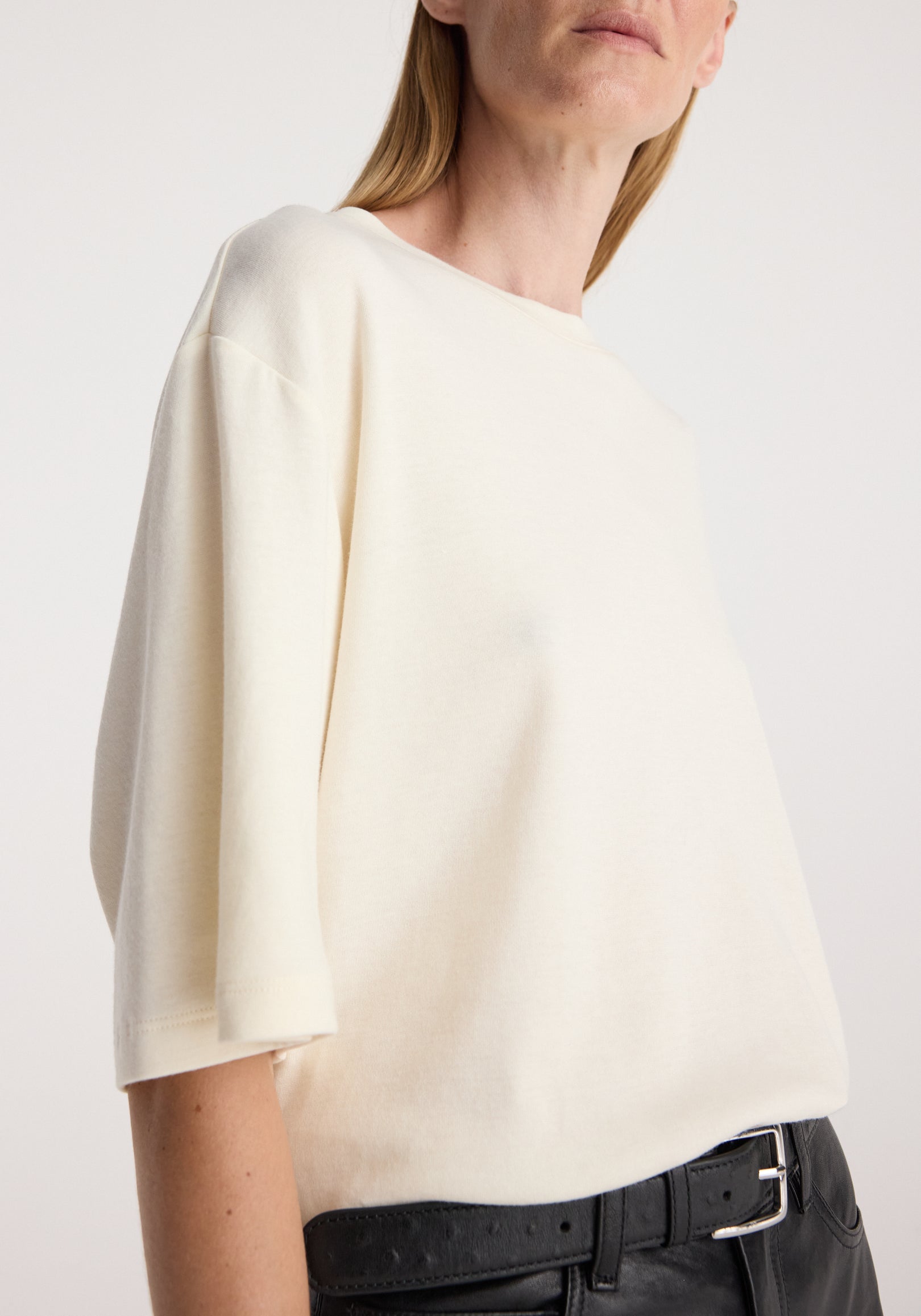 Woman wearing an oversized cotton wool t-shirt | cream in a relaxed fit with half sleeves, paired with black pants and a belt against a plain background.