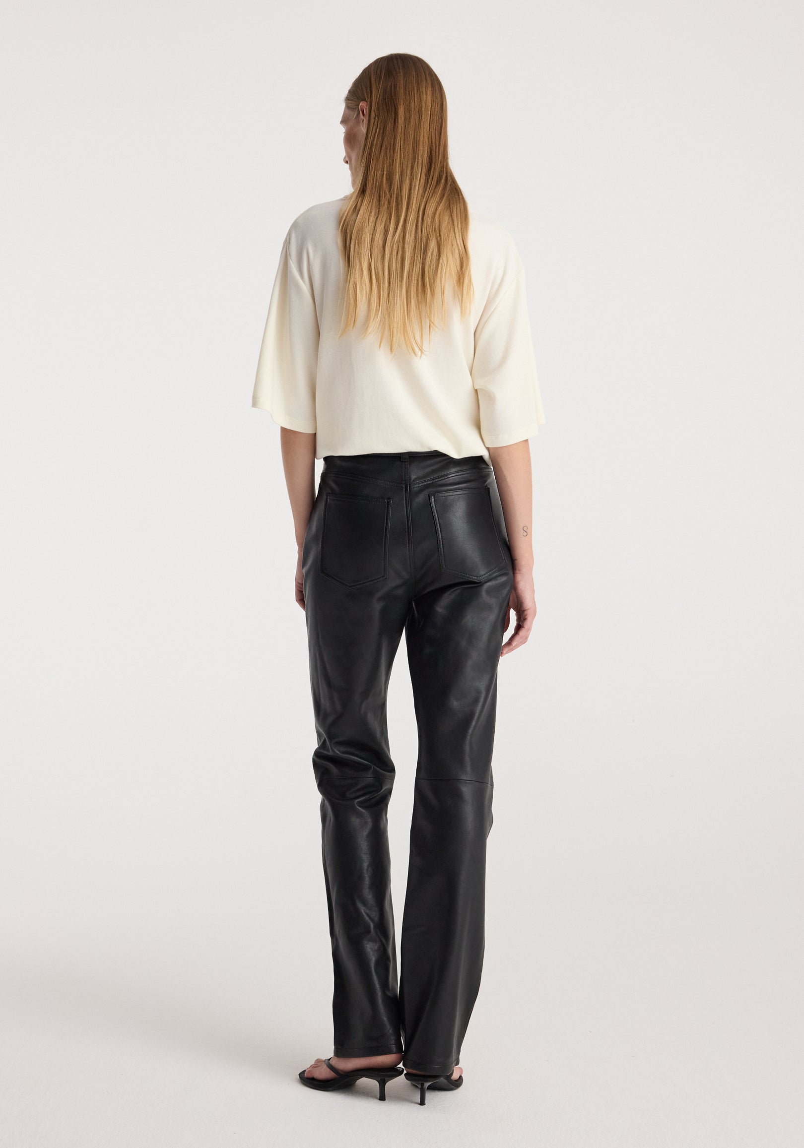 Woman with long hair wearing an oversized cotton wool t-shirt | cream, paired with black leather pants and black heeled sandals, shown from the back against a white background.