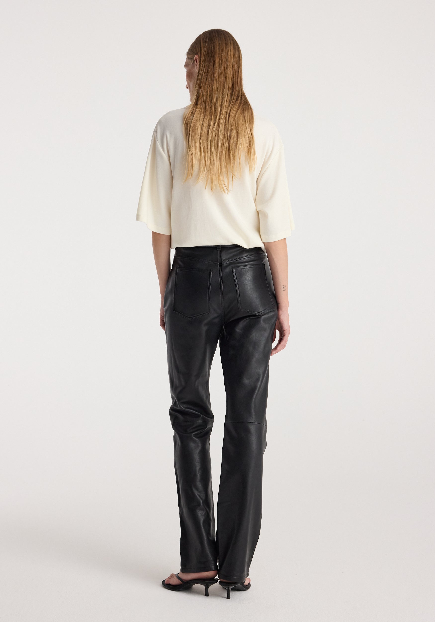 Woman with long hair wearing an oversized cotton wool t-shirt | cream, paired with black leather pants and black heeled sandals, shown from the back against a white background.