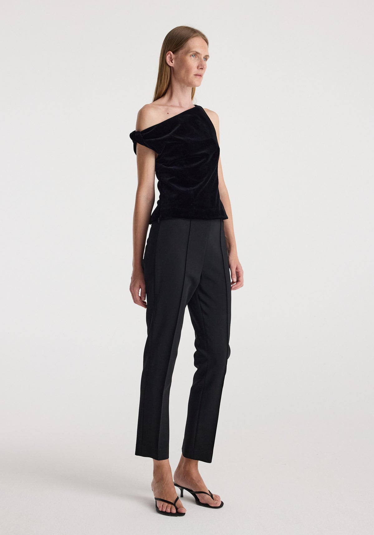 Woman wearing a draped velvet top | black with an asymmetric off-shoulder design paired with black tailored pants and black heeled sandals against a plain background.