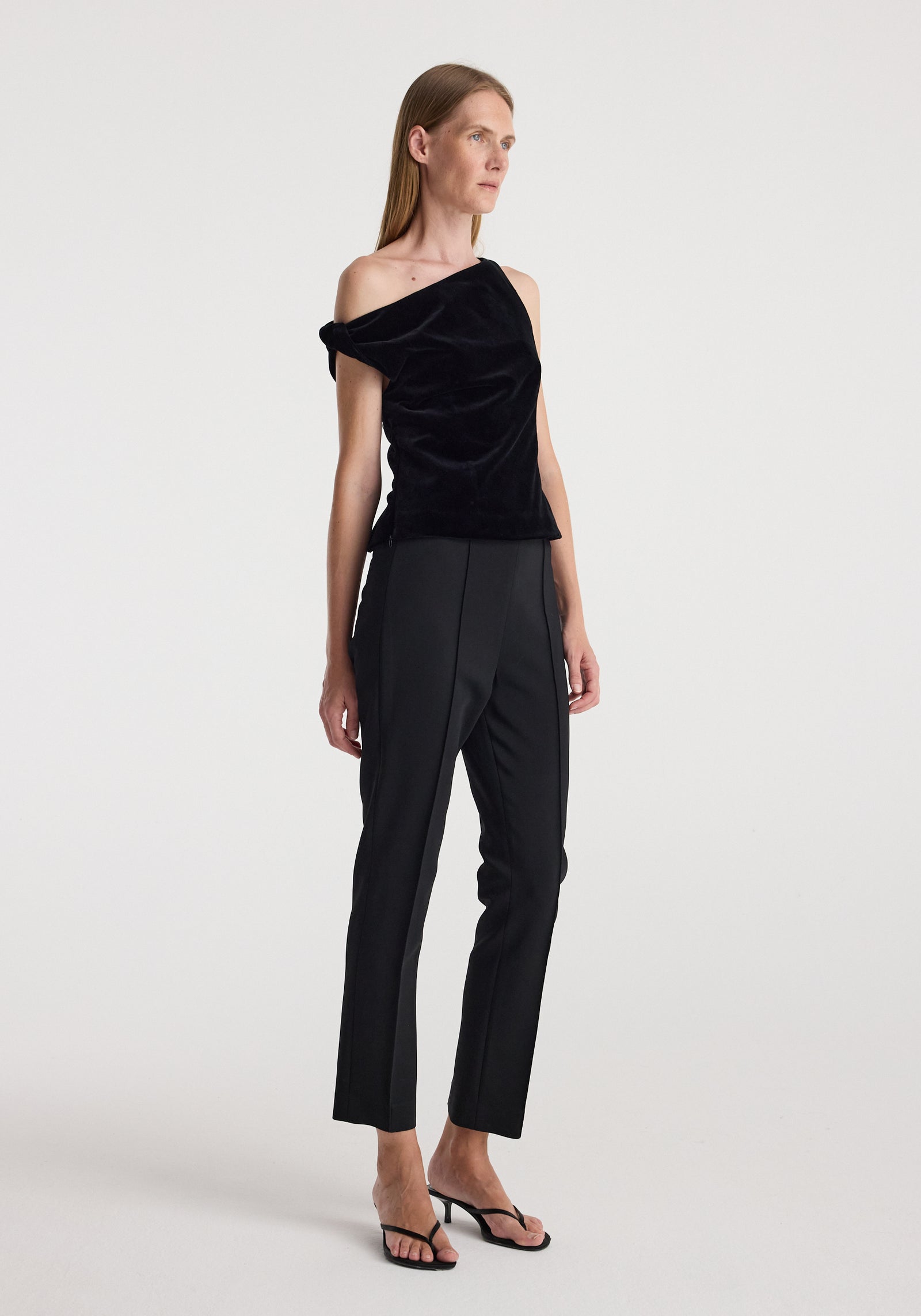 Woman wearing a draped velvet top | black with an asymmetric off-shoulder design paired with black tailored pants and black heeled sandals against a plain background.
