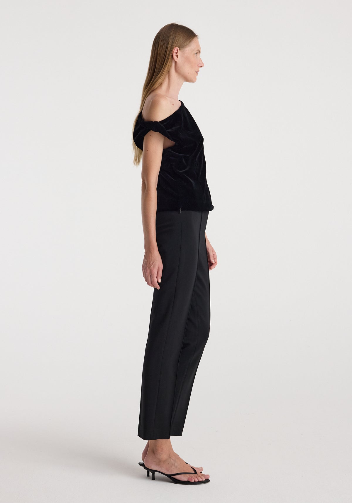 Woman standing in profile wearing a black draped velvet top | black with off-shoulder detail, paired with black tailored pants and black heeled sandals against a plain background.