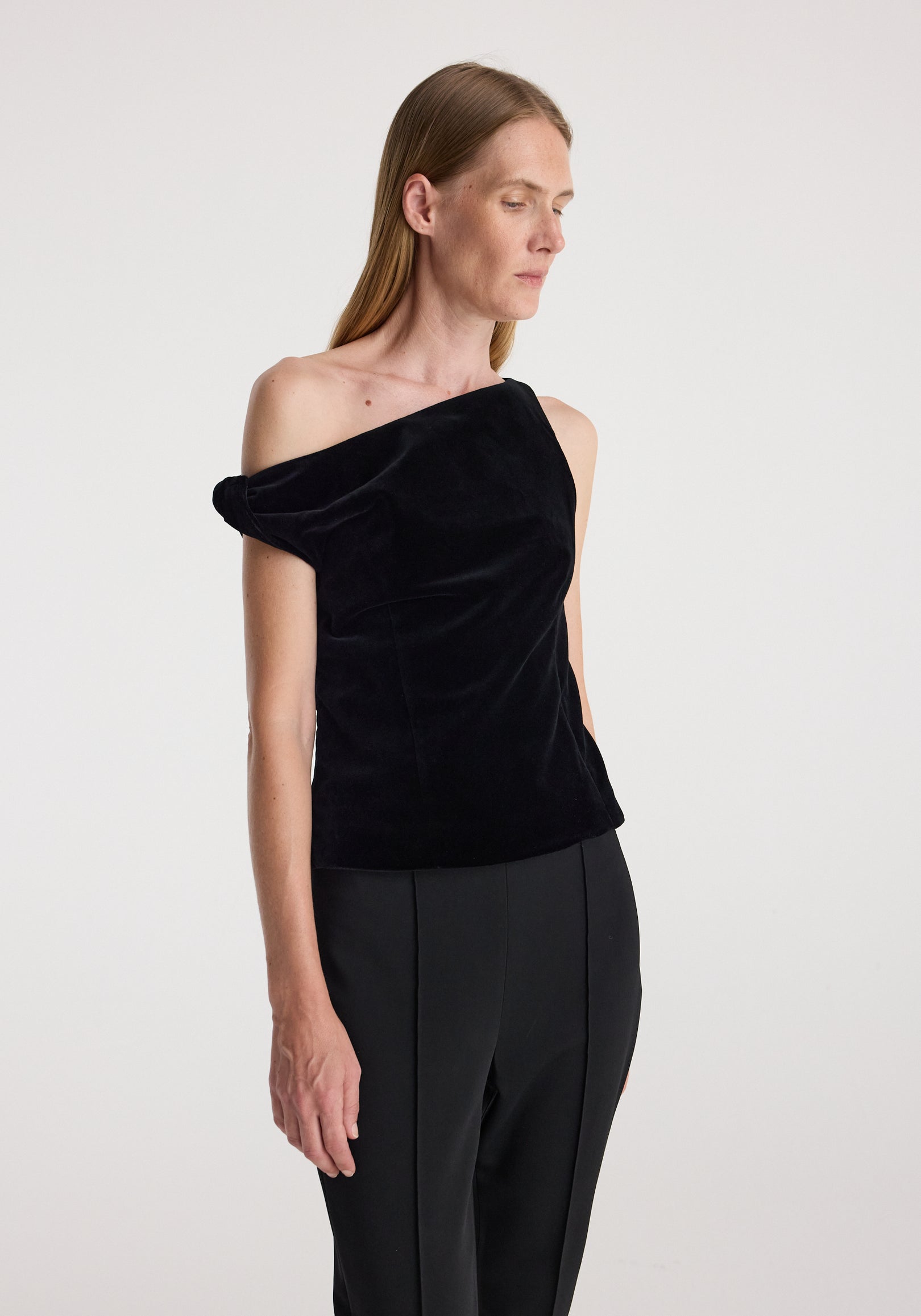 Woman wearing a draped velvet top | black with an asymmetrical neckline and soft texture paired with black tailored pants against a plain background.