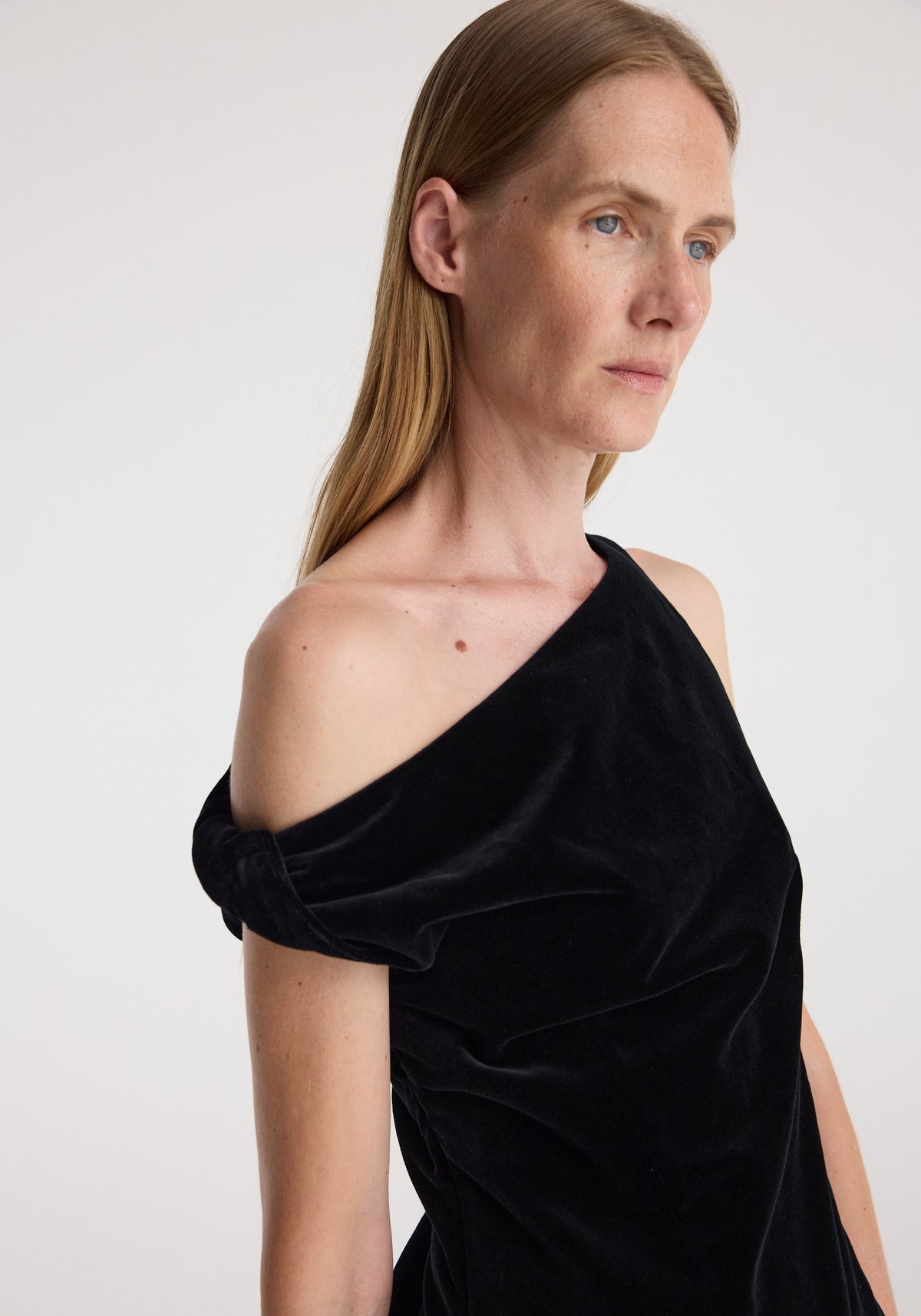 Close-up of a woman wearing a draped velvet top | black with a one-shoulder design and soft texture against a plain light background.