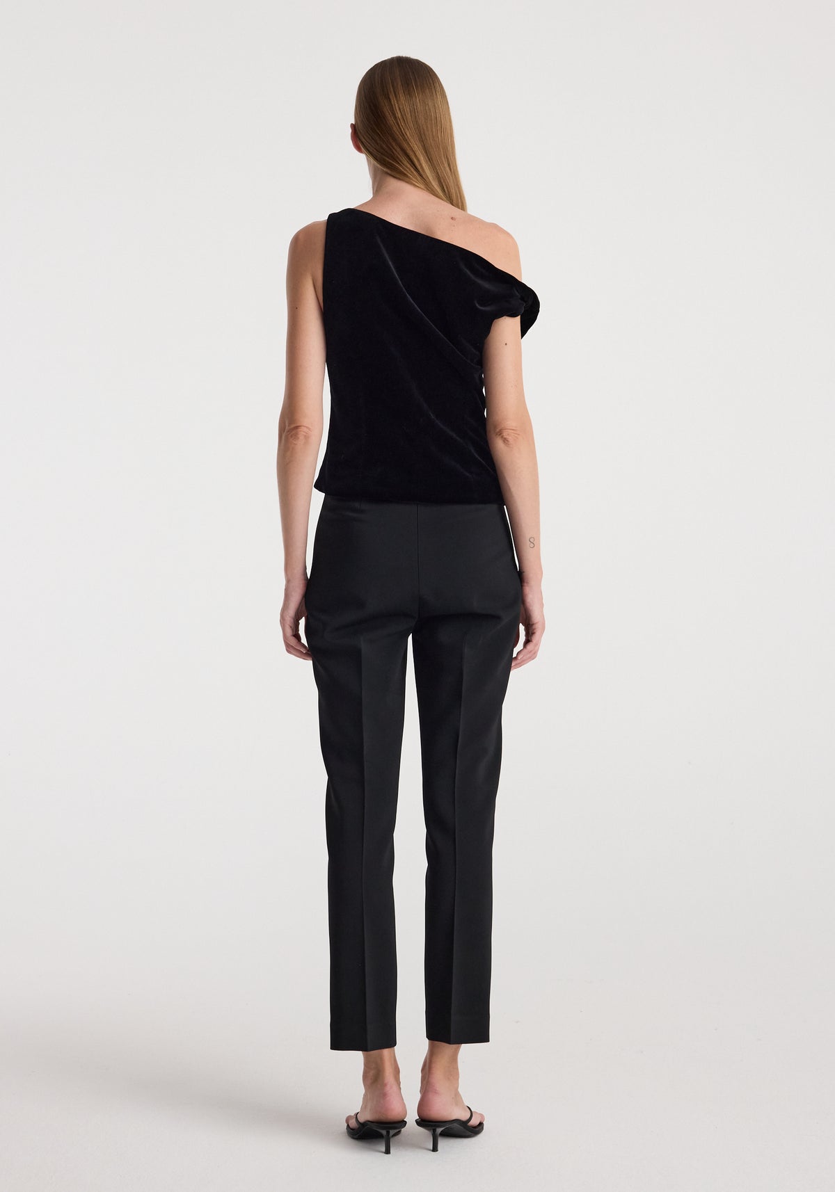 Woman seen from the back wearing a black Draped velvet top | black with one shoulder exposed, paired with black fitted pants and black heeled sandals against a plain white background.
