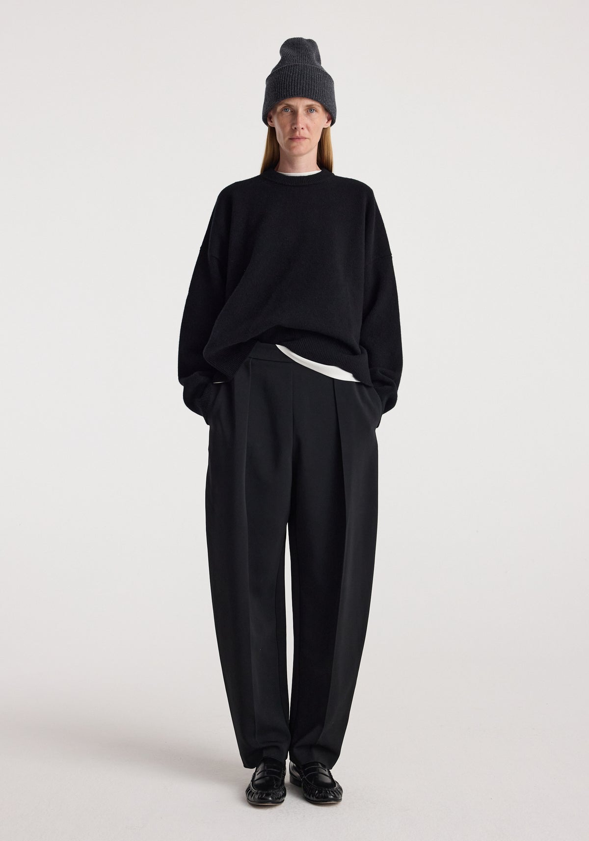 Woman wearing a dark grey knitted Beanie | anthracite melange, paired with a black sweater, black wide-leg trousers, and black shoes against a plain background.