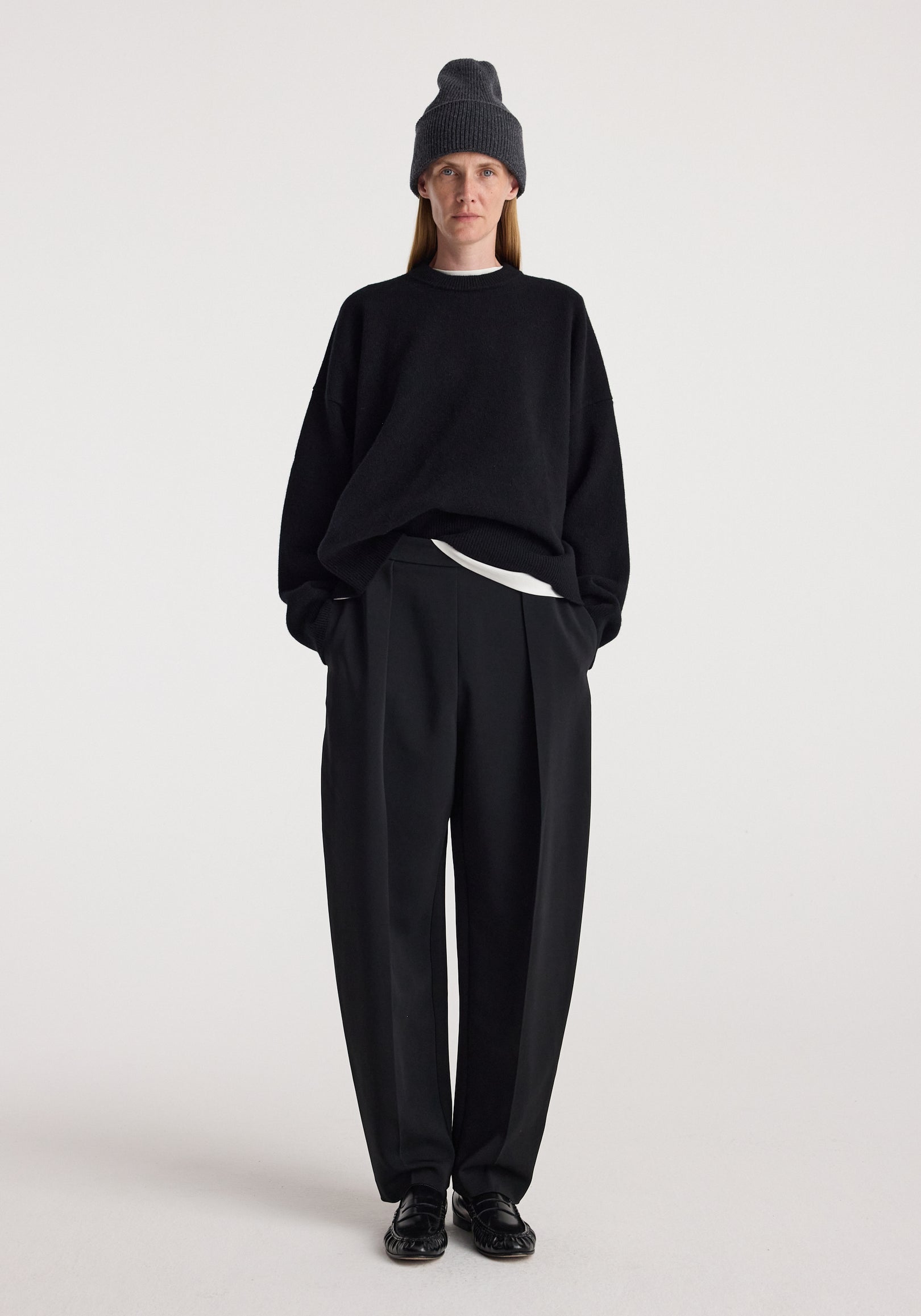 Woman wearing a dark grey knitted Beanie | anthracite melange, paired with a black sweater, black wide-leg trousers, and black shoes against a plain background.