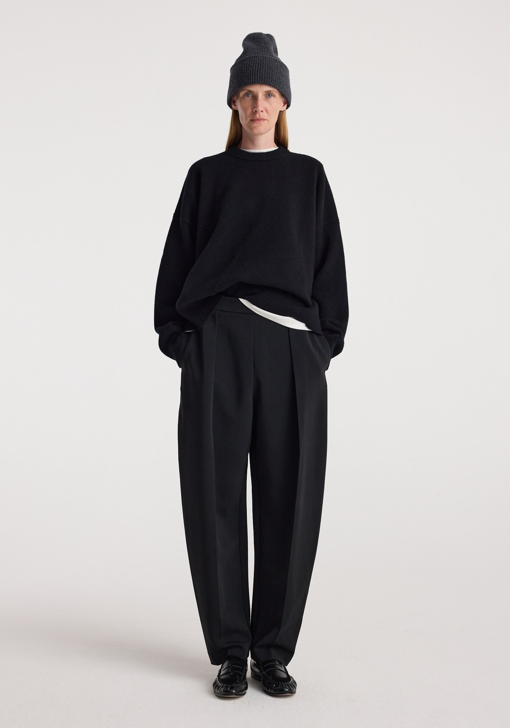 Woman wearing a dark grey knitted Beanie | anthracite melange, paired with a black sweater, black wide-leg trousers, and black shoes against a plain background.