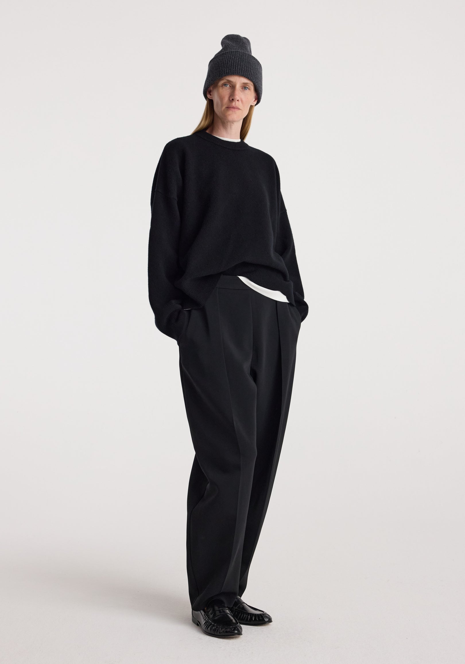 Woman wearing a dark grey knitted Beanie | anthracite melange with casual black sweater, black wide-leg pants, and black shoes standing against a plain white background.