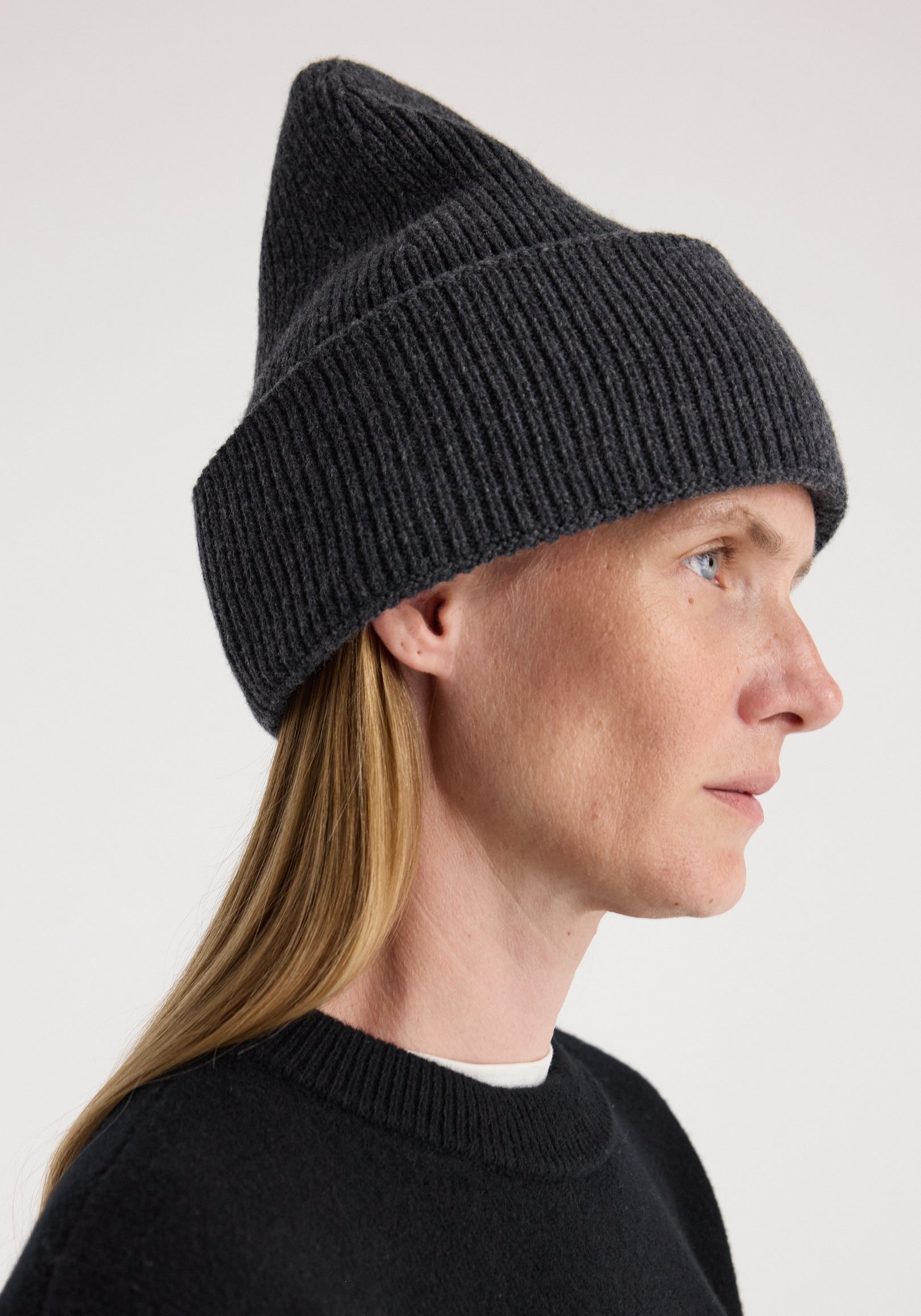 Woman with long blonde hair wearing a dark knit hat styled with a pointed top, titled Beanie | anthracite melange, paired with a black crew neck sweater against a neutral background.