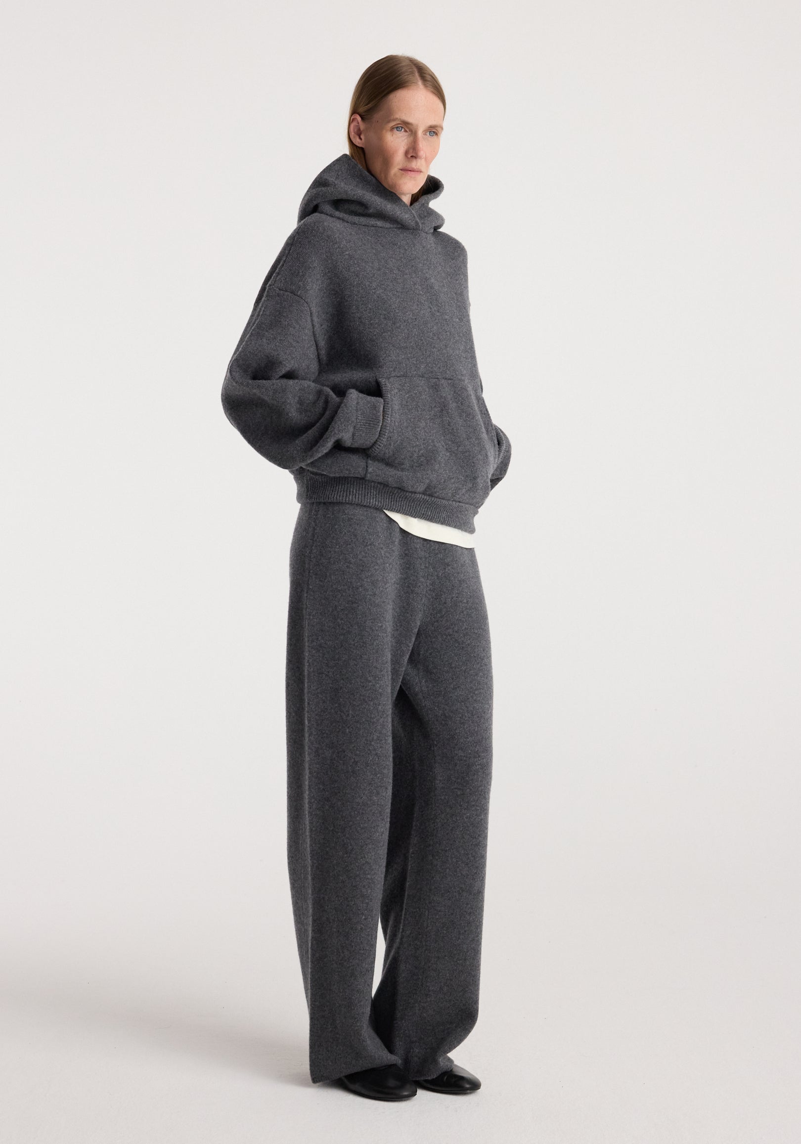 Person standing in a relaxed pose wearing dark grey melange cozy knitwear including Compact knitted trousers | dark grey melange, paired with a matching oversized hoodie.