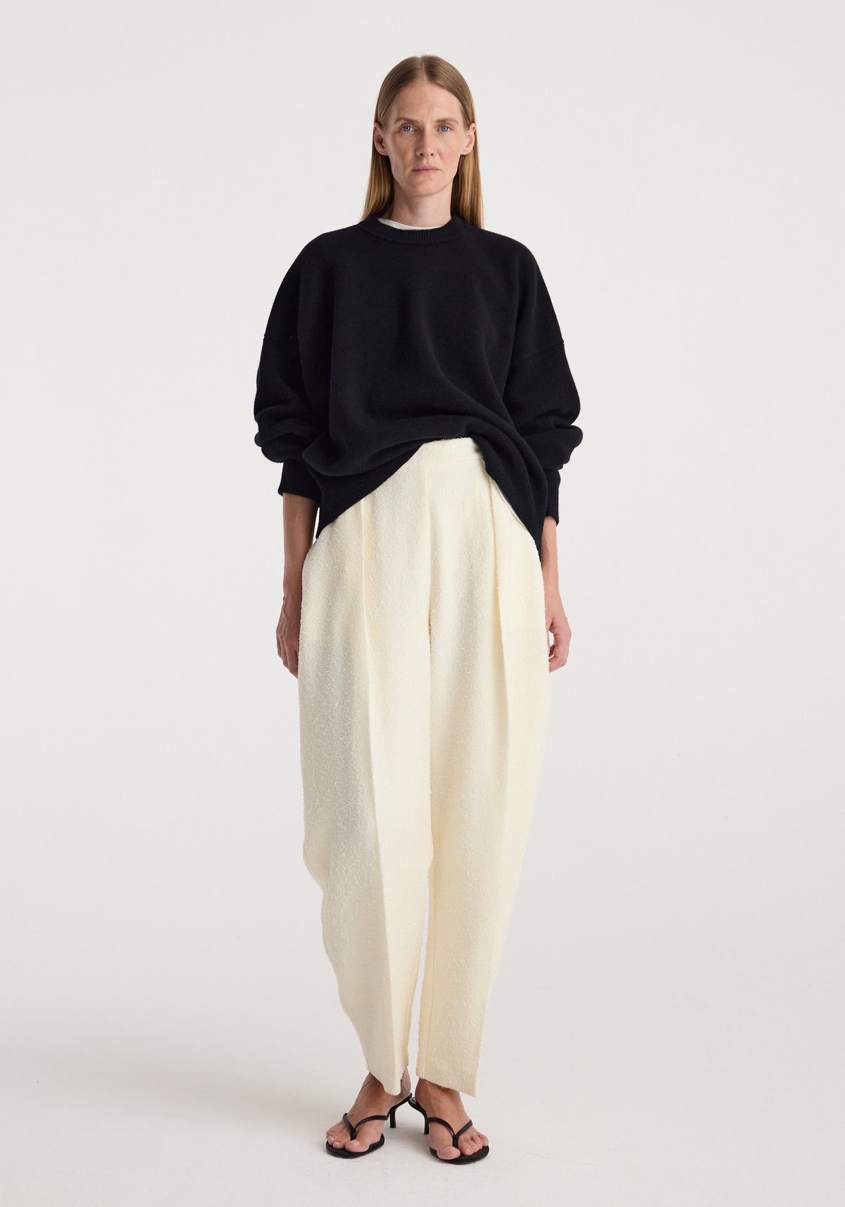 Model wearing a Classic crewneck jumper | black paired with loose white trousers and black open-toe heels against a plain light background.
