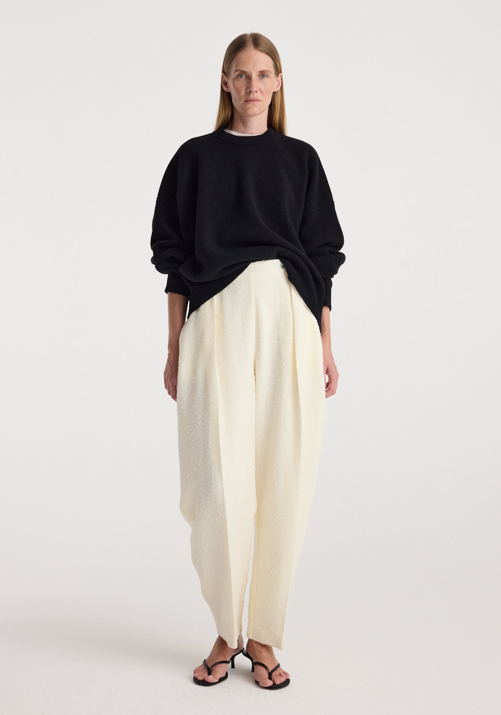 Model wearing a Classic crewneck jumper | black paired with loose white trousers and black open-toe heels against a plain light background.