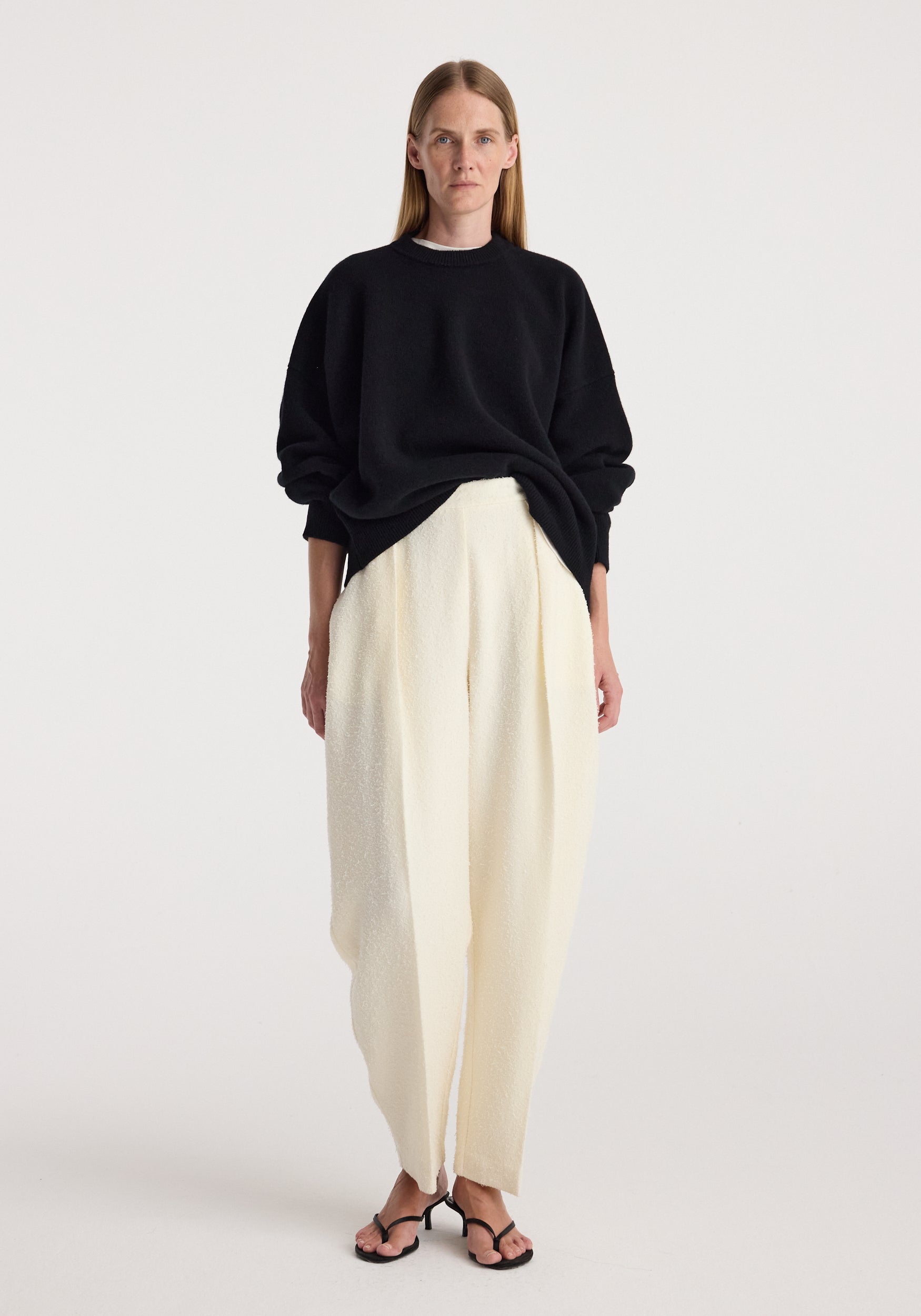 Model wearing a Classic crewneck jumper | black paired with loose white trousers and black open-toe heels against a plain light background.