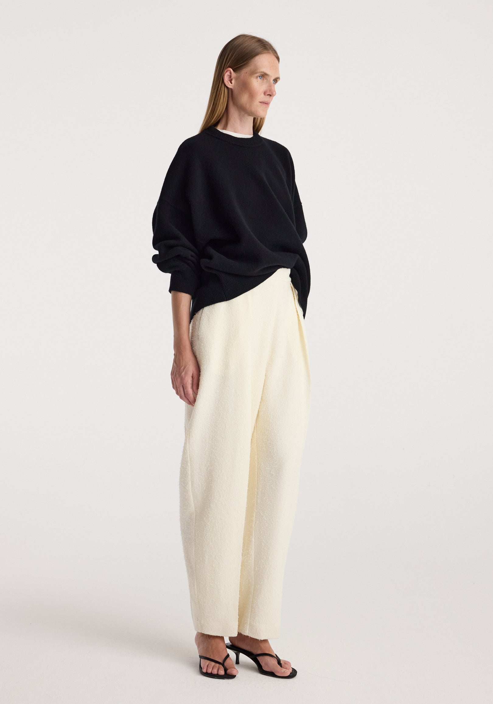 Woman modeling a loose-fitting black Classic crewneck jumper | black paired with white wide-leg pants and black open-toe heels against a plain background.