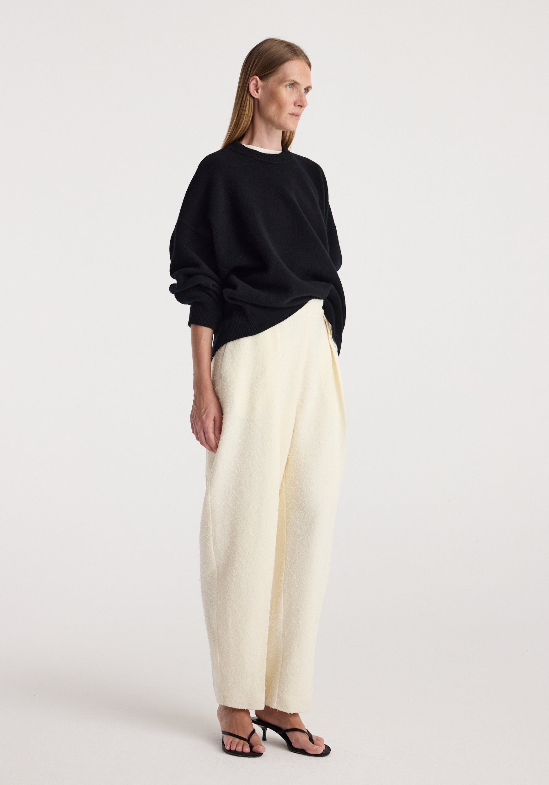 Woman modeling a loose-fitting black Classic crewneck jumper | black paired with white wide-leg pants and black open-toe heels against a plain background.
