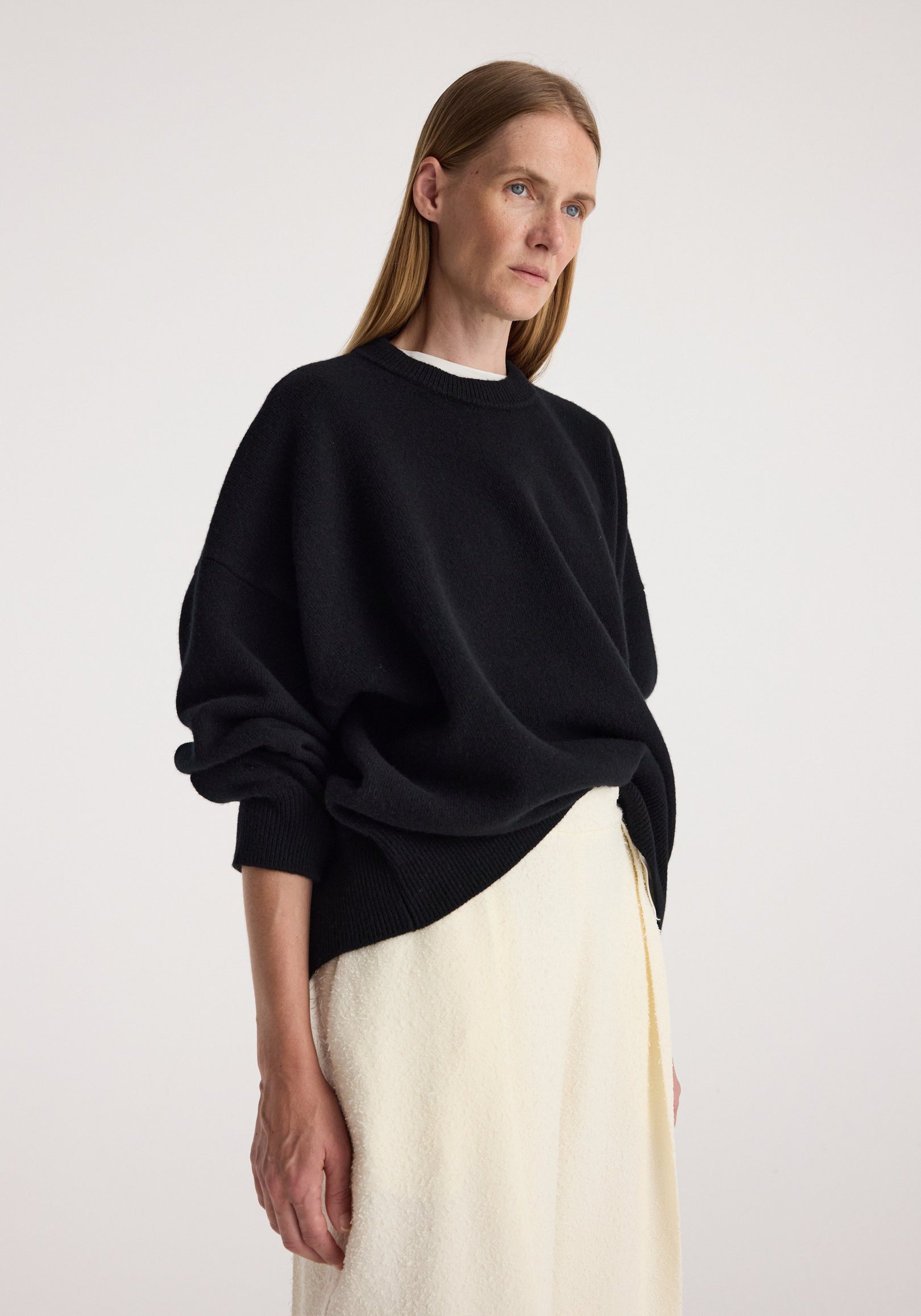 Woman wearing a relaxed fit Classic crewneck jumper | black with ribbed cuffs and hem, paired with a light cream skirt against a plain background.