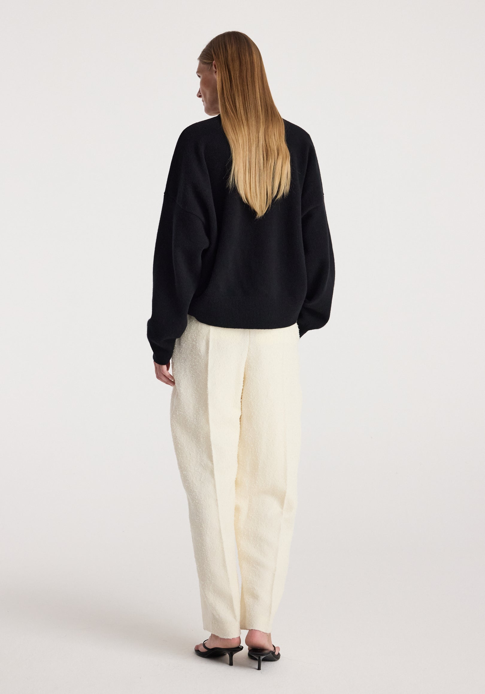 Woman wearing a black Classic crewneck jumper | black paired with loose-fitting cream-colored pants and black heeled sandals, viewed from the back against a plain background.