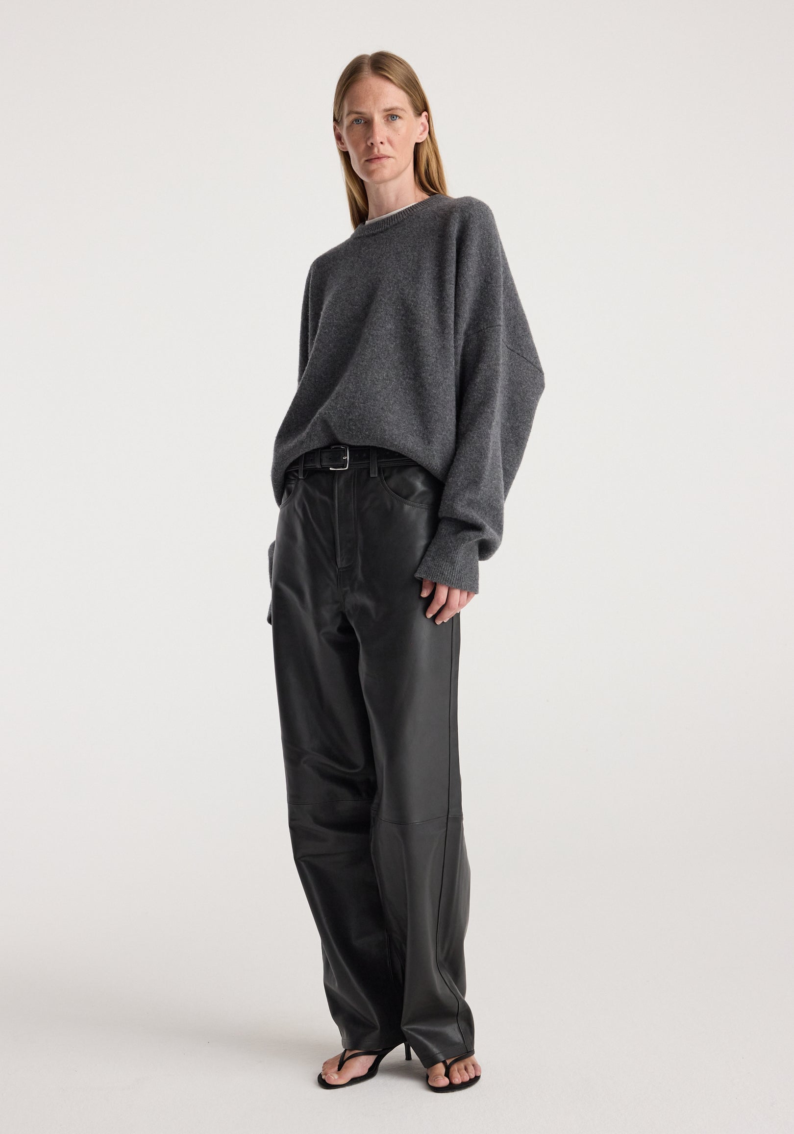Woman wearing a Classic crewneck jumper | dark grey melange with loose fit, paired with black leather pants and black open-toe heels, standing against a plain background.