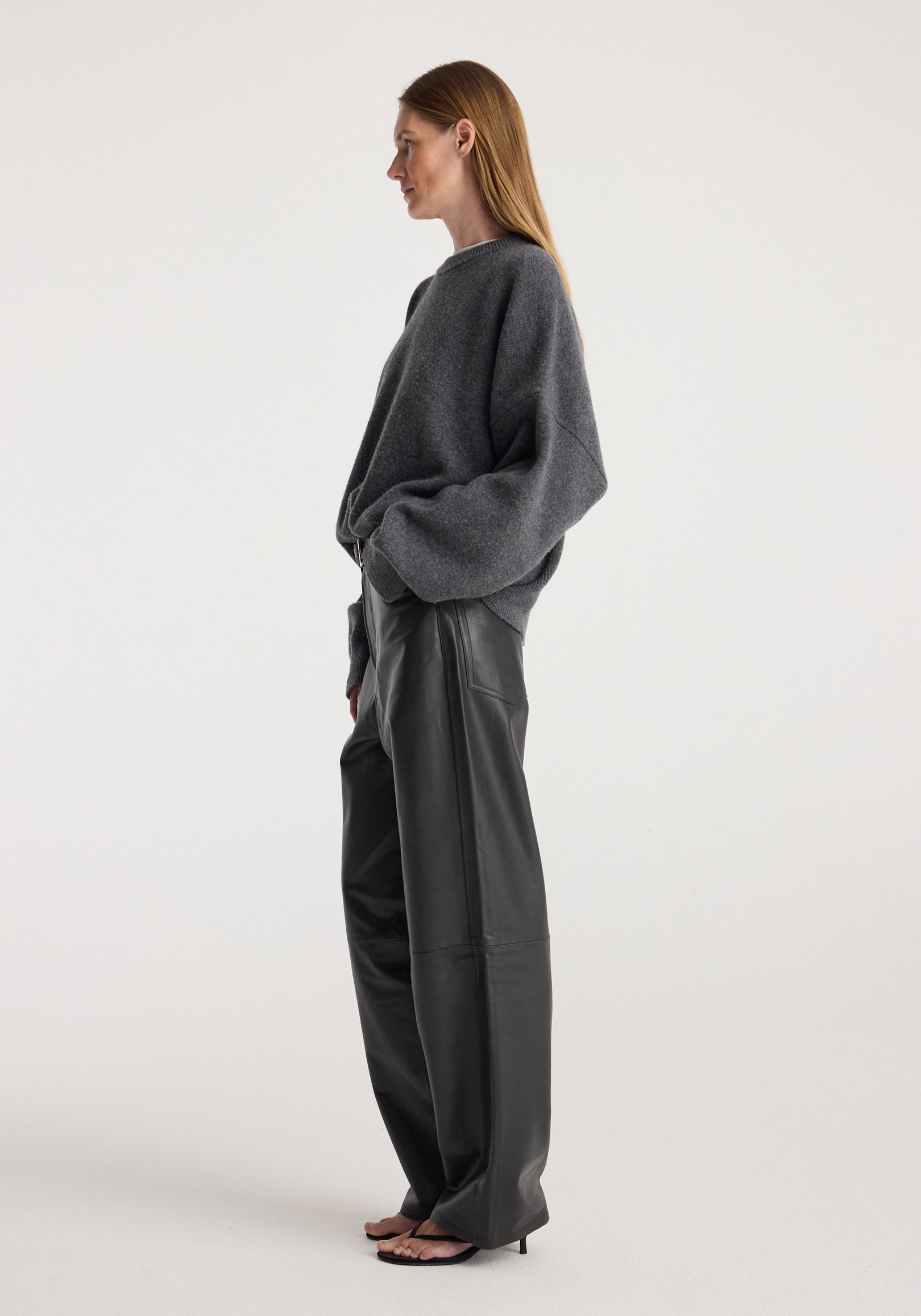 Woman wearing a Classic crewneck jumper | dark grey melange with long sleeves, paired with black wide-leg leather pants and black heeled sandals, standing in profile.
