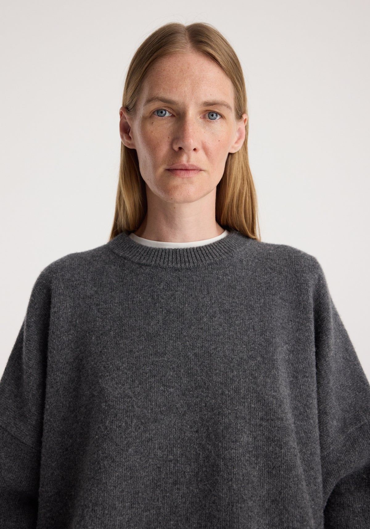 Woman wearing a soft classic crewneck jumper | dark grey melange with a relaxed fit and ribbed neckline, styled over a white underlayer against a plain background.