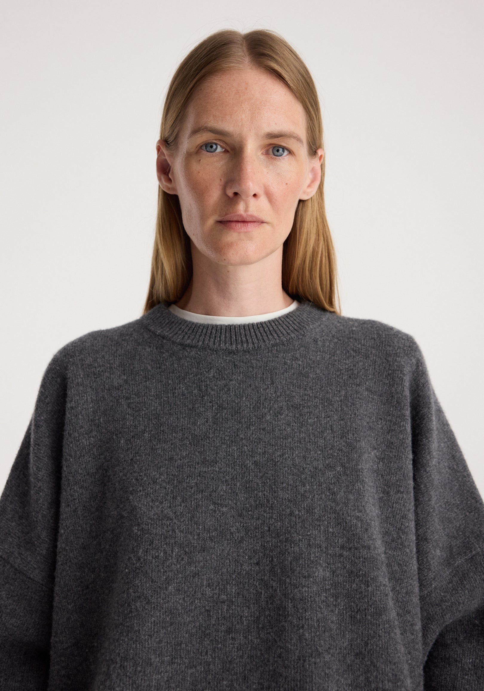 Woman wearing a soft classic crewneck jumper | dark grey melange with a relaxed fit and ribbed neckline, styled over a white underlayer against a plain background.