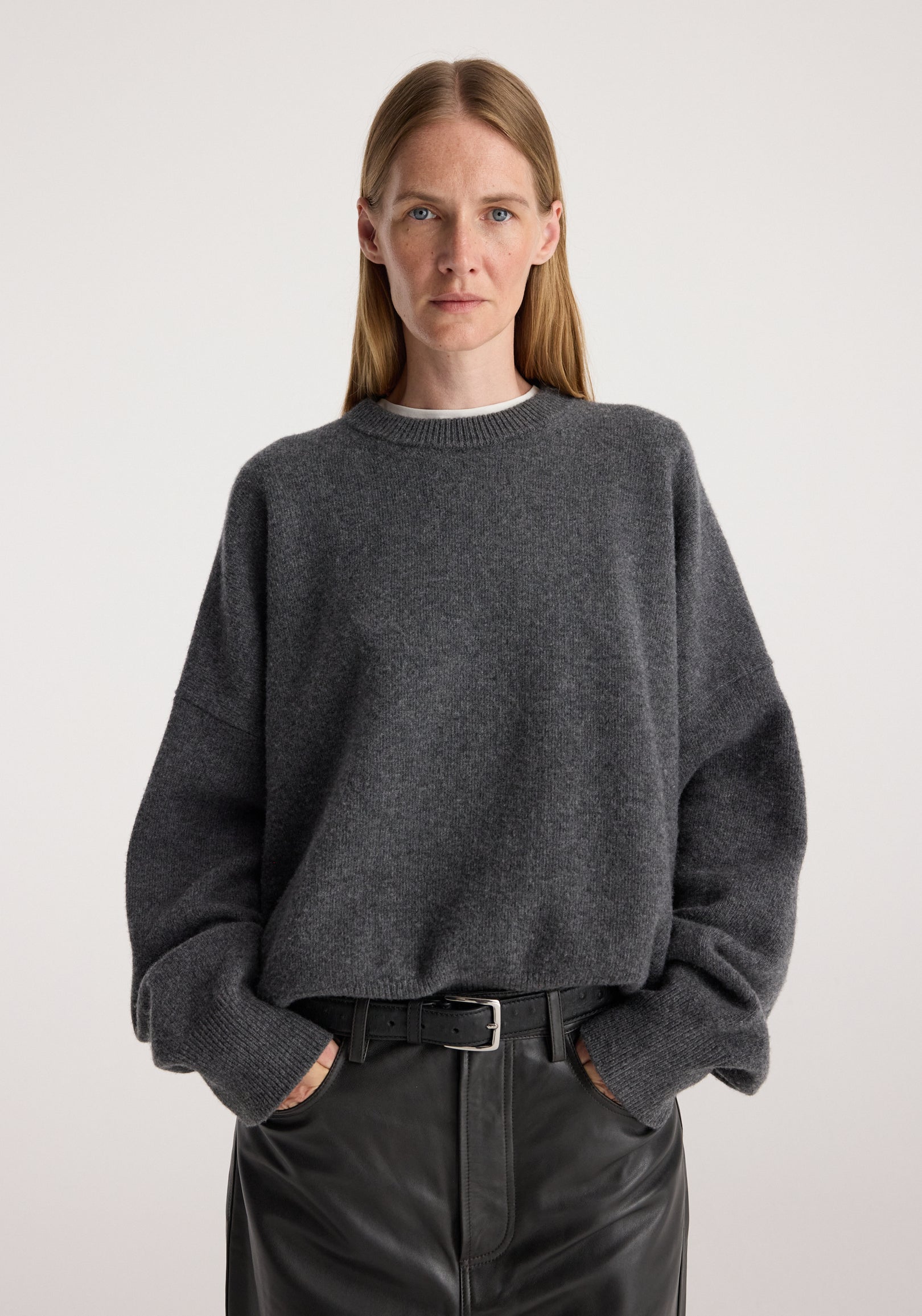 Woman wearing a dark grey melange Classic crewneck jumper with long sleeves and relaxed fit, tucked into black leather pants with a black belt.