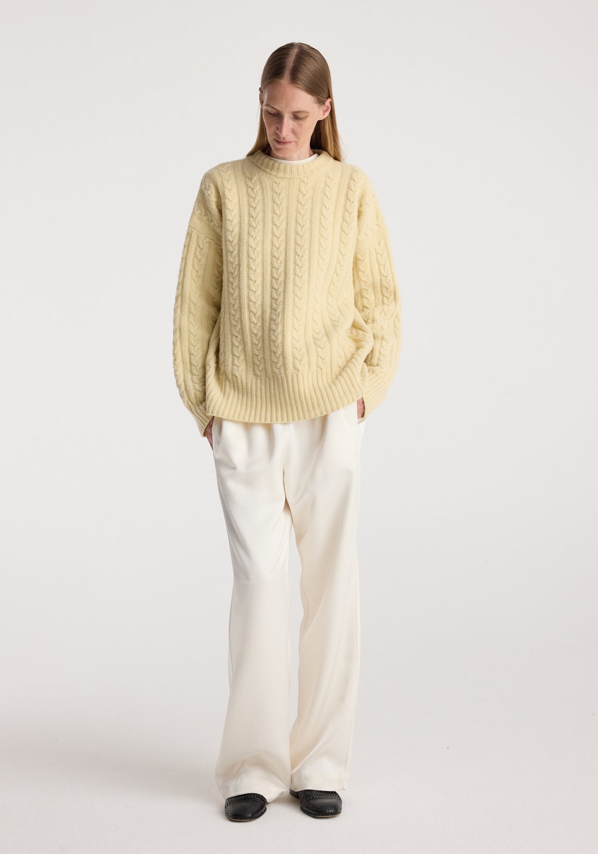 Woman wearing a butter-colored Cable knit jumper | butter with long sleeves paired with loose white trousers and black shoes, standing against a plain background.