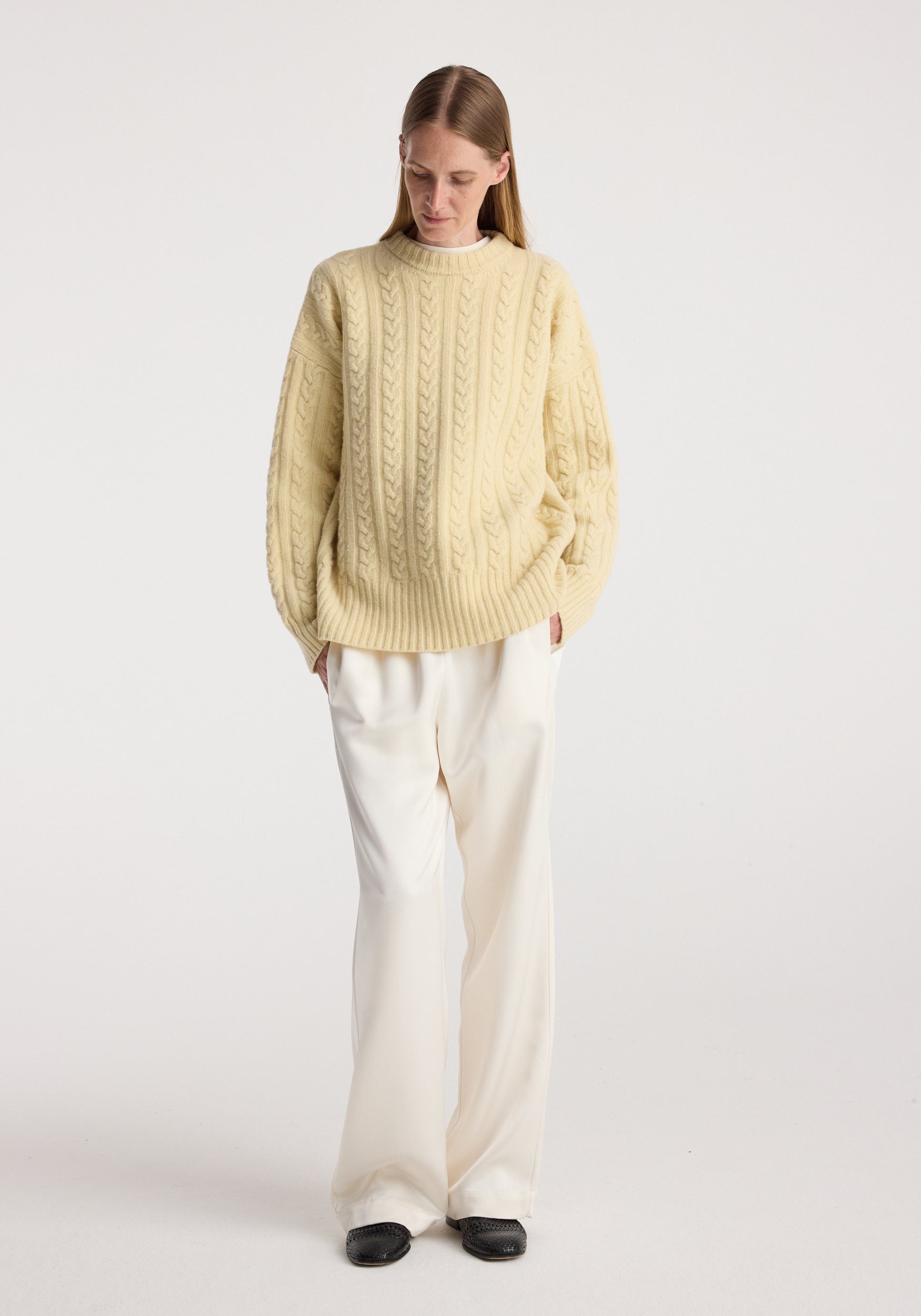 Woman wearing a butter-colored Cable knit jumper | butter with long sleeves paired with loose white trousers and black shoes, standing against a plain background.