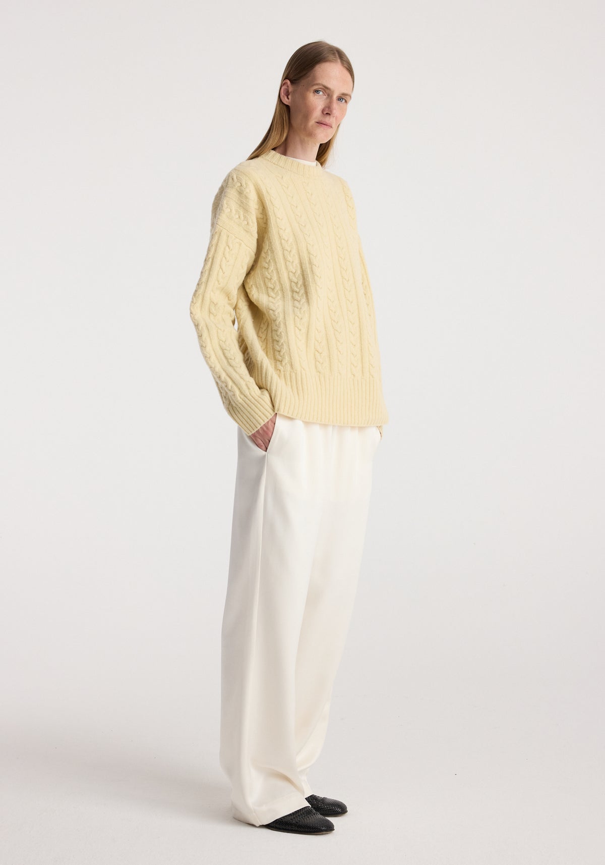 Woman wearing a light yellow cable knit jumper | butter with long sleeves, paired with loose white trousers and black shoes, standing against a plain background.
