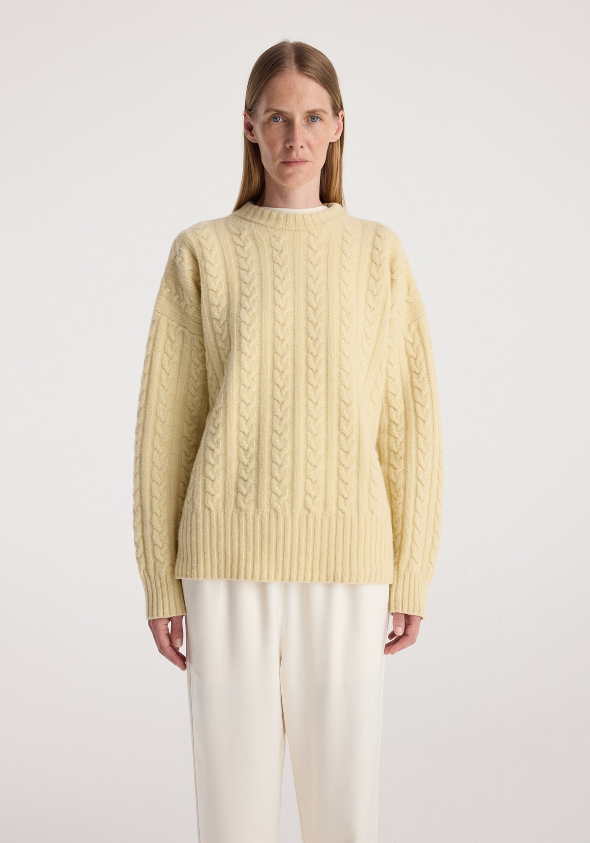 Woman wearing a cream-colored cable knit jumper | butter with long sleeves and ribbed hem, paired with loose white trousers, standing against a plain background.