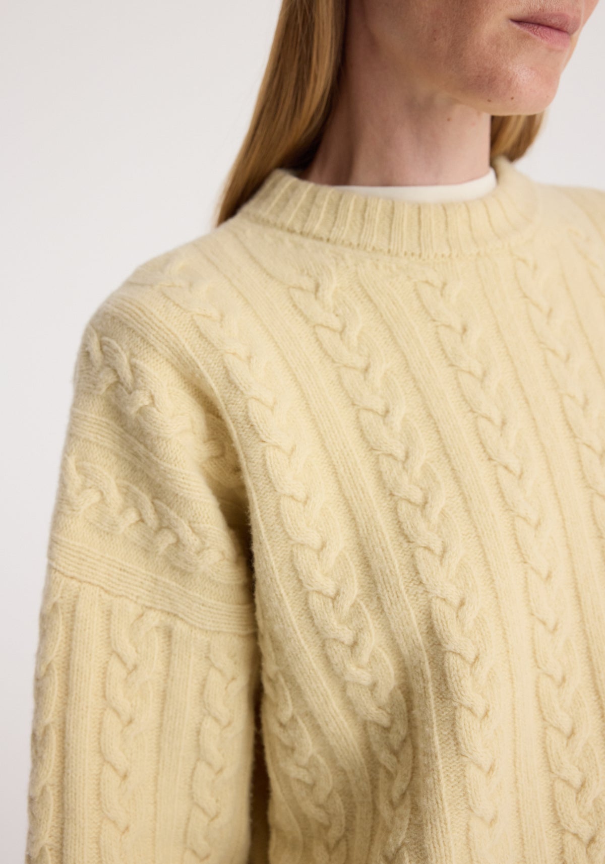 Close-up of a person wearing a soft, cream-colored Cable knit jumper | butter with detailed braided knit patterns and a ribbed crew neckline.