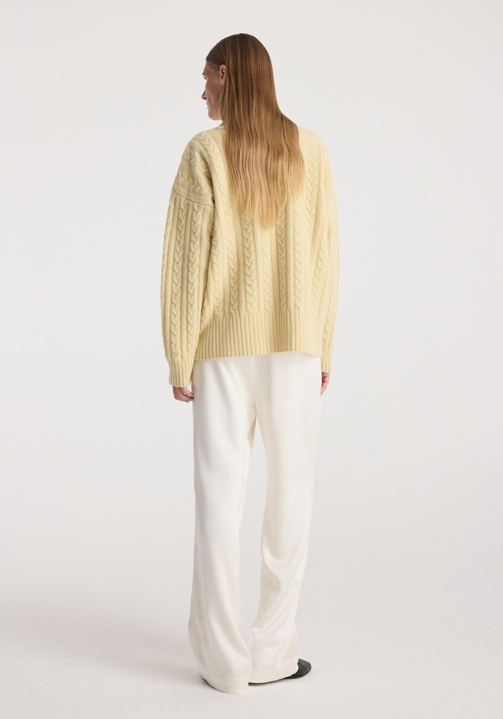 Woman seen from behind wearing a butter-colored cable knit jumper | butter with long sleeves paired with loose white pants against a plain background.