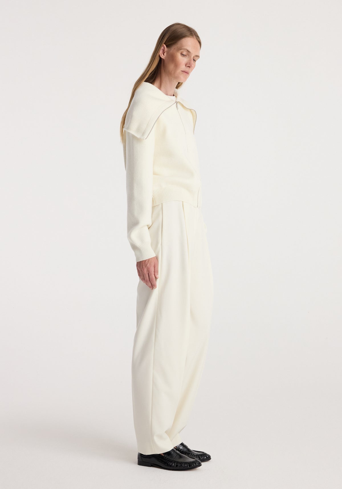 Woman wearing a cream-colored Two way sailor zip cardigan | cream with wide-leg trousers and black shoes, standing in profile against a plain background.