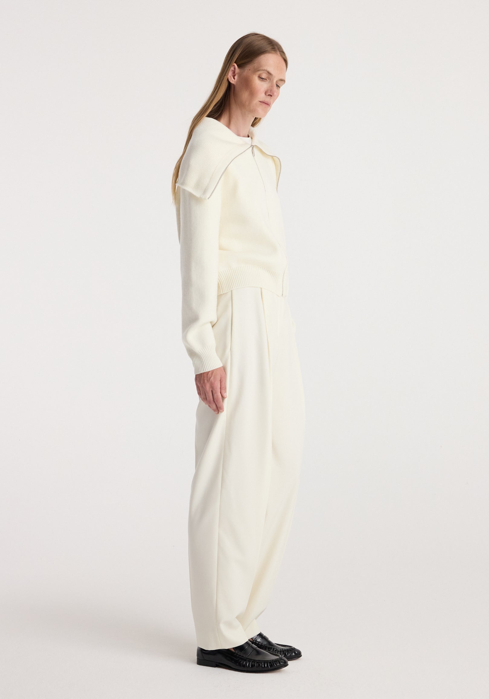 Woman wearing a cream-colored Two way sailor zip cardigan | cream with wide-leg trousers and black shoes, standing in profile against a plain background.