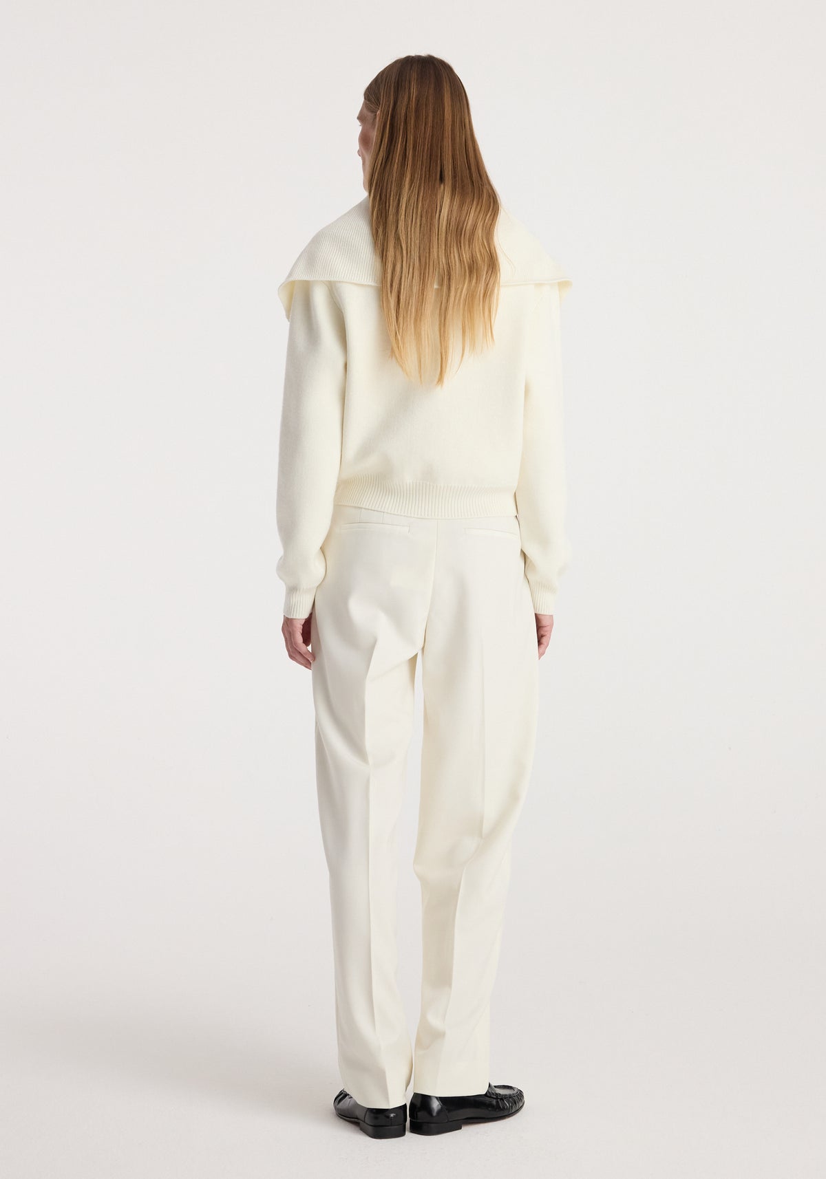 Woman seen from the back wearing a cream-colored Two way sailor zip cardigan | cream with matching cream trousers and black shoes against a plain white background.