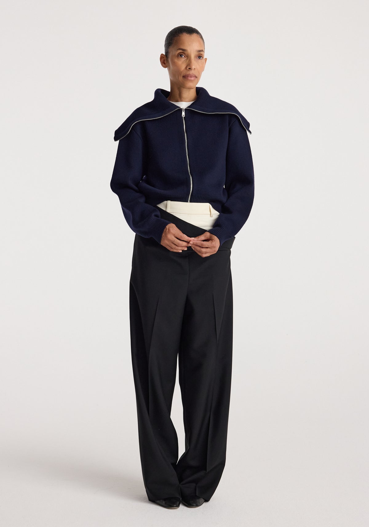 Female model wearing a navy Two way sailor zip cardigan | navy with a large collar and long sleeves, paired with black high-waisted wide-leg trousers.