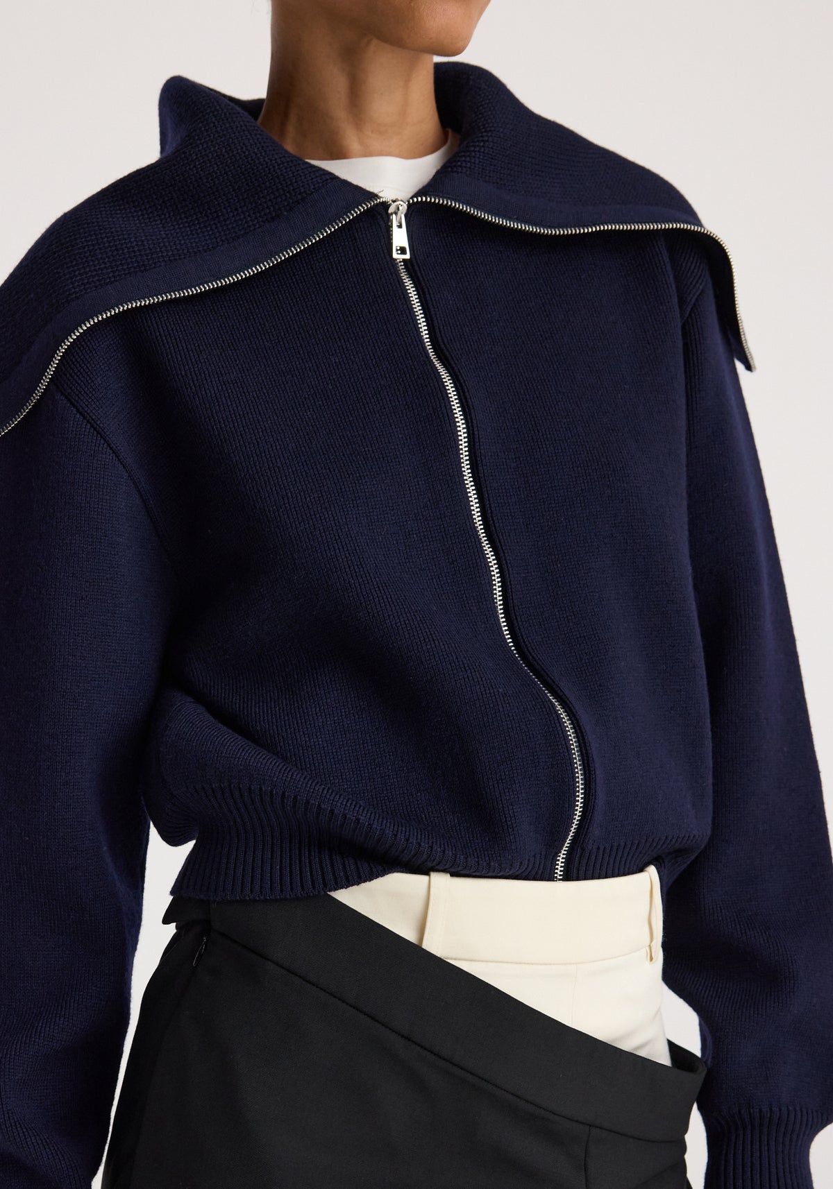Close-up of a person wearing a navy blue Two way sailor zip cardigan | navy with a silver zipper partially unzipped, paired with light beige and black pants.