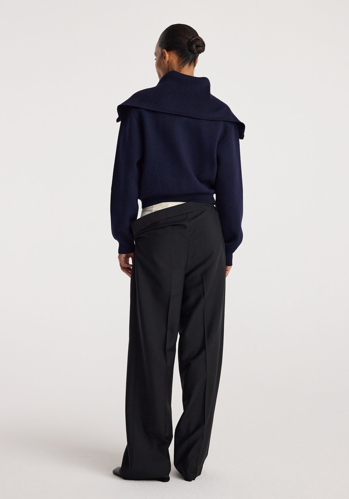 Back view of a person wearing a navy Two way sailor zip cardigan with a large collar paired with loose black trousers against a plain background.
