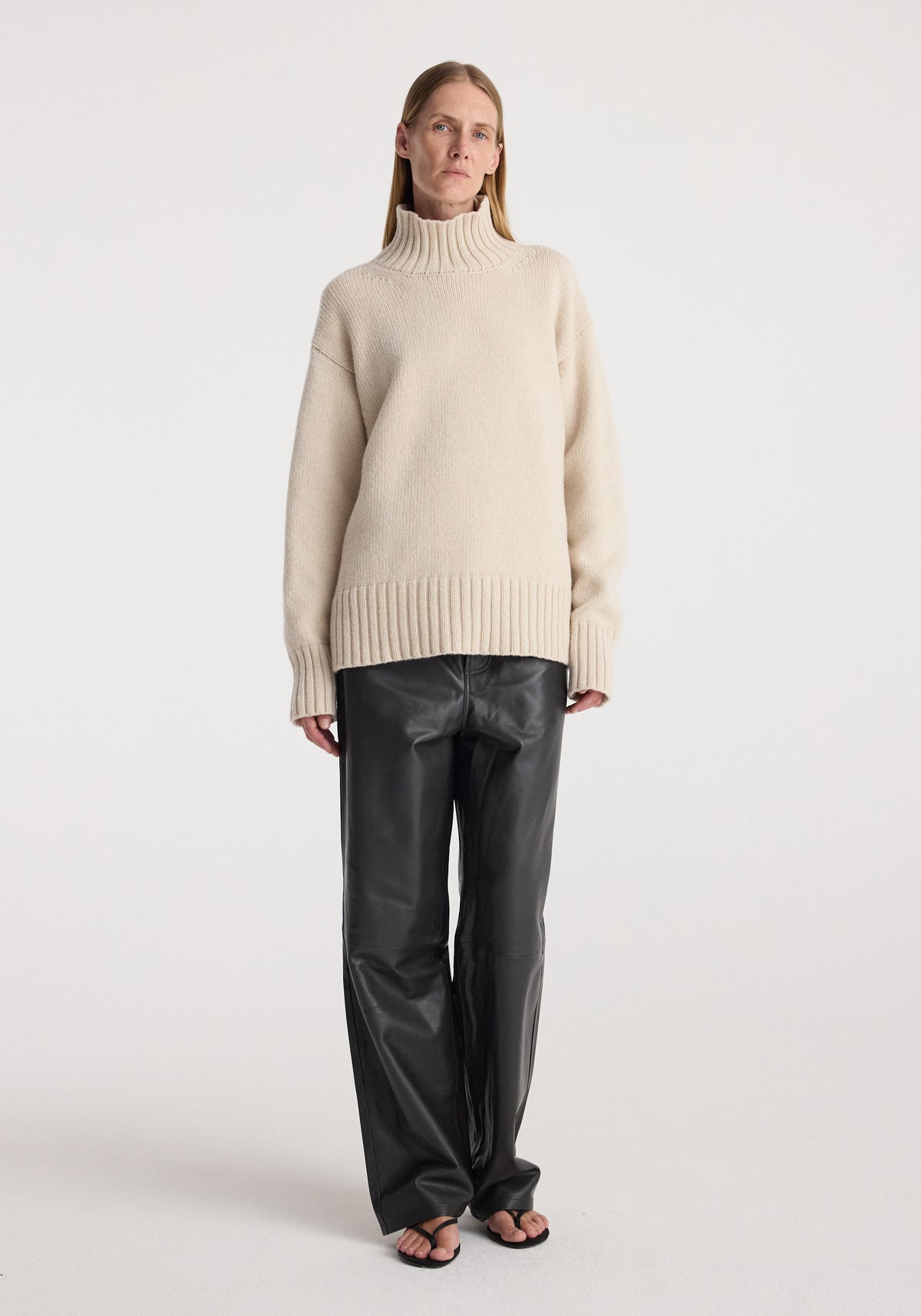 Woman wearing a Wool-cashmere turtleneck | oat with long sleeves and ribbed cuffs, paired with black leather wide-leg pants and open-toe sandals, standing against a plain background.