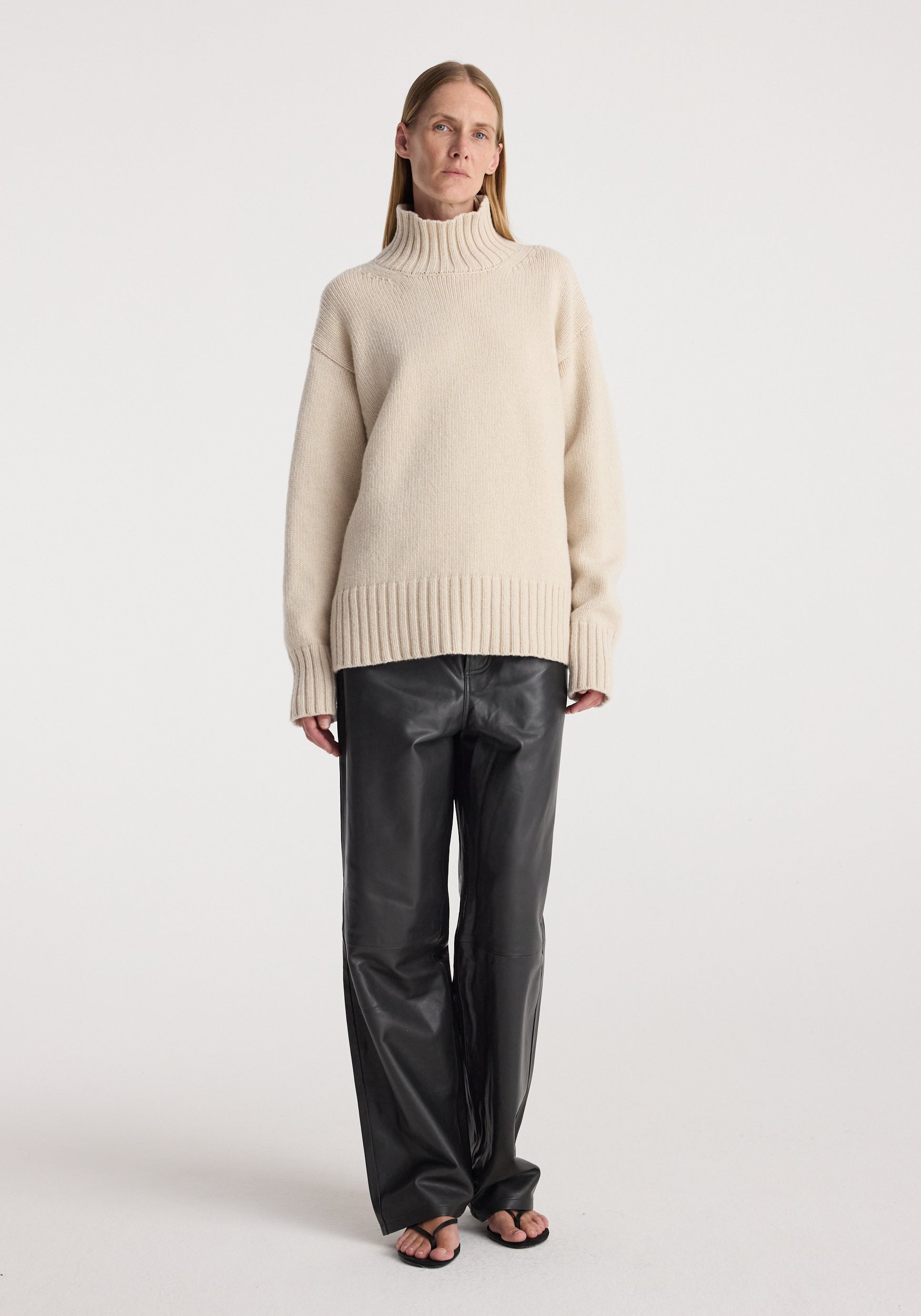 Woman wearing a Wool-cashmere turtleneck | oat with long sleeves and ribbed cuffs, paired with black leather wide-leg pants and open-toe sandals, standing against a plain background.