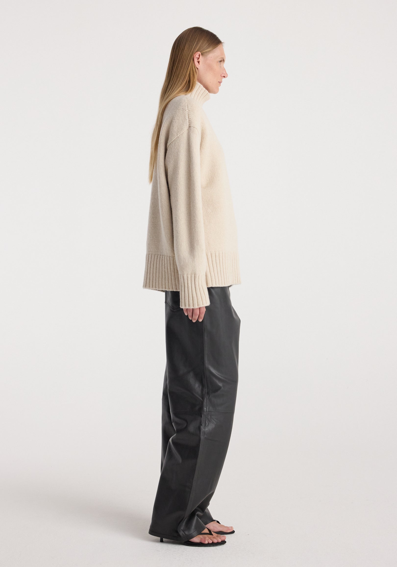 Profile view of a woman wearing a Wool-cashmere turtleneck | oat paired with loose black pants and open-toe heels against a plain white background.