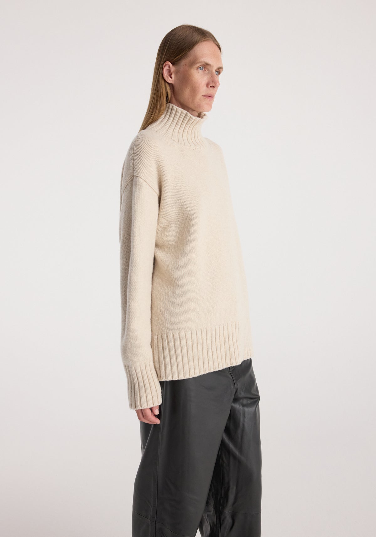 Side view of a person wearing a Wool-cashmere turtleneck | oat in a soft beige color, paired with black pants, against a plain light background.