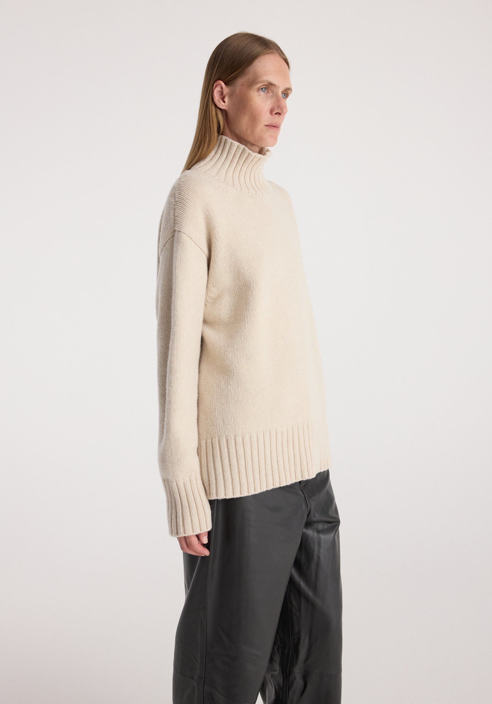 Side view of a person wearing a Wool-cashmere turtleneck | oat in a soft beige color, paired with black pants, against a plain light background.