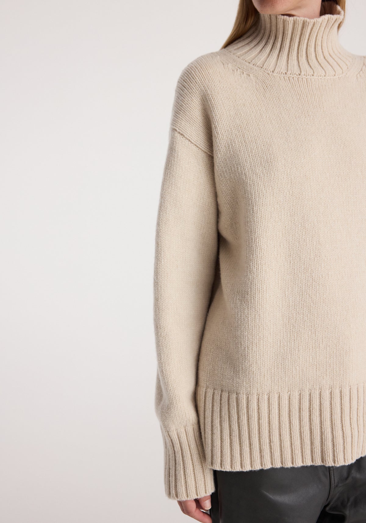 Close-up of a person wearing a soft, light oat-colored Wool-cashmere turtleneck | oat sweater with ribbed cuffs, hem, and high neck, paired with dark pants.