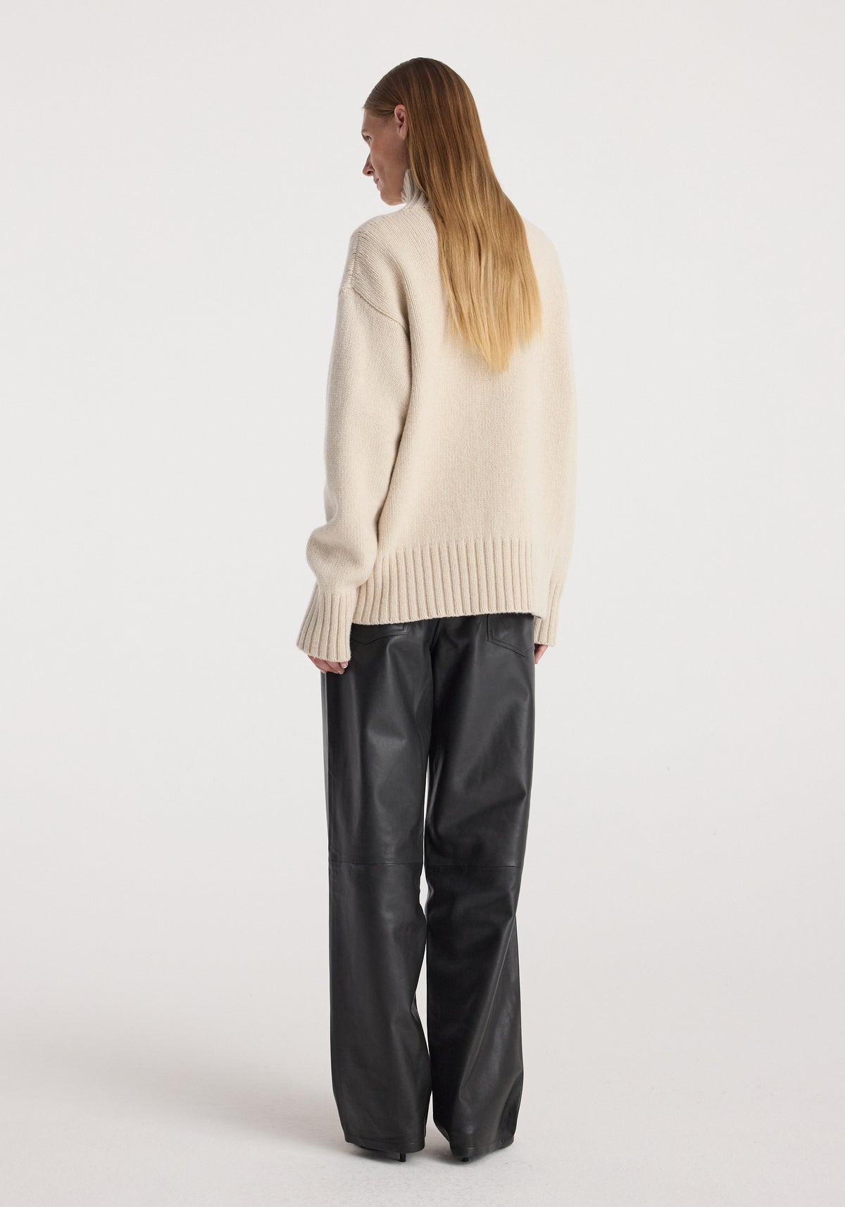 Woman wearing a Wool-cashmere turtleneck | oat in a soft beige color, paired with loose black leather pants, photographed from behind against a plain background.